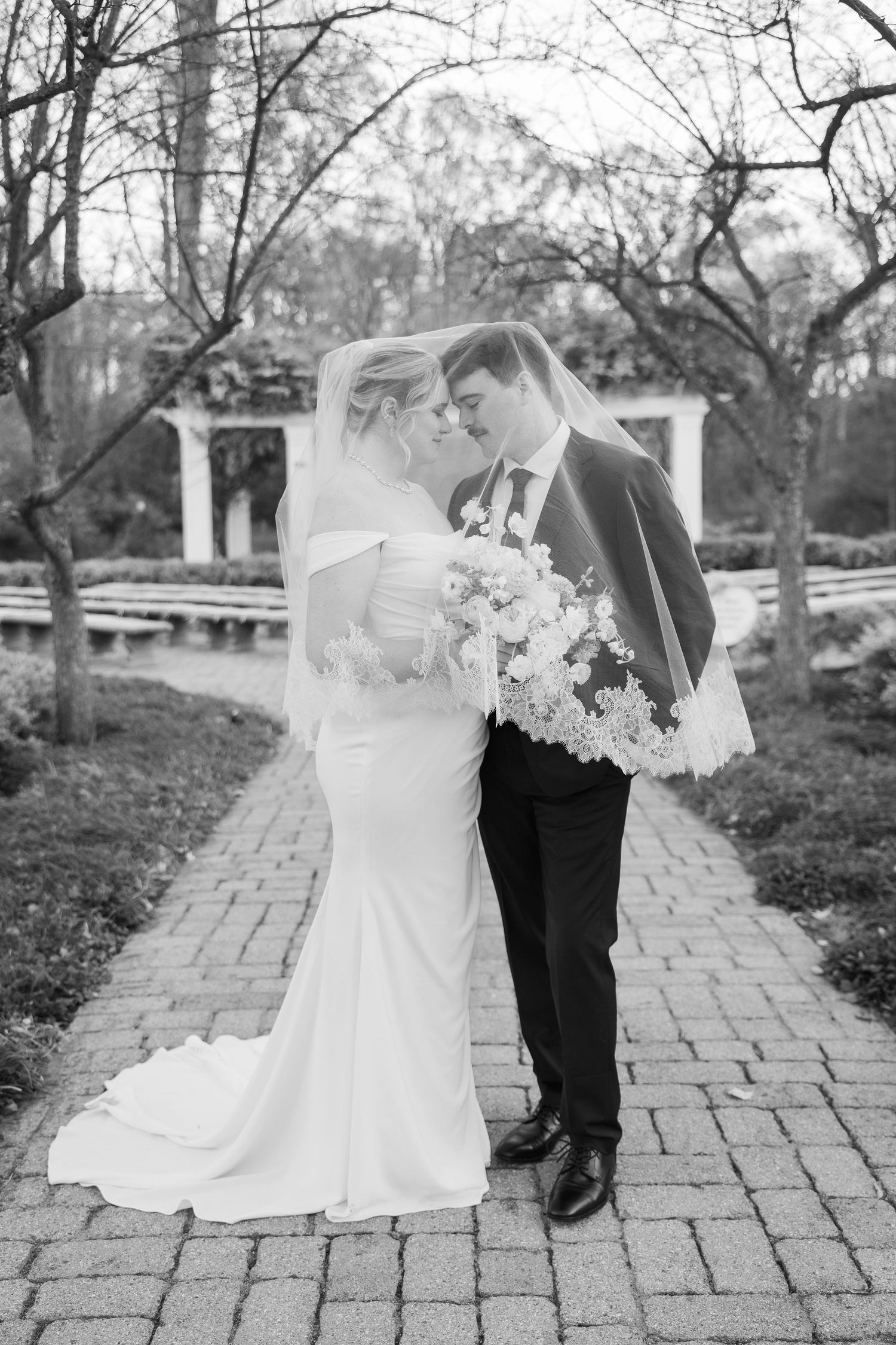 Romantic wedding portrait on brick path with white gazebo and garden backdrop.