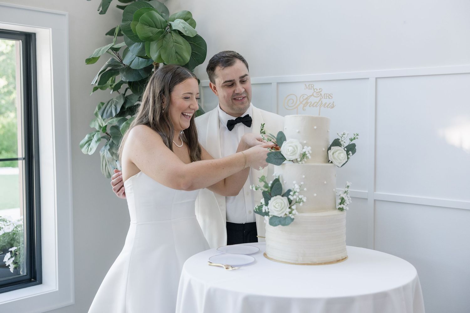 Newlyweds smiling as they cut their elegant white tiered wedding cake adorned with white roses and greenery.