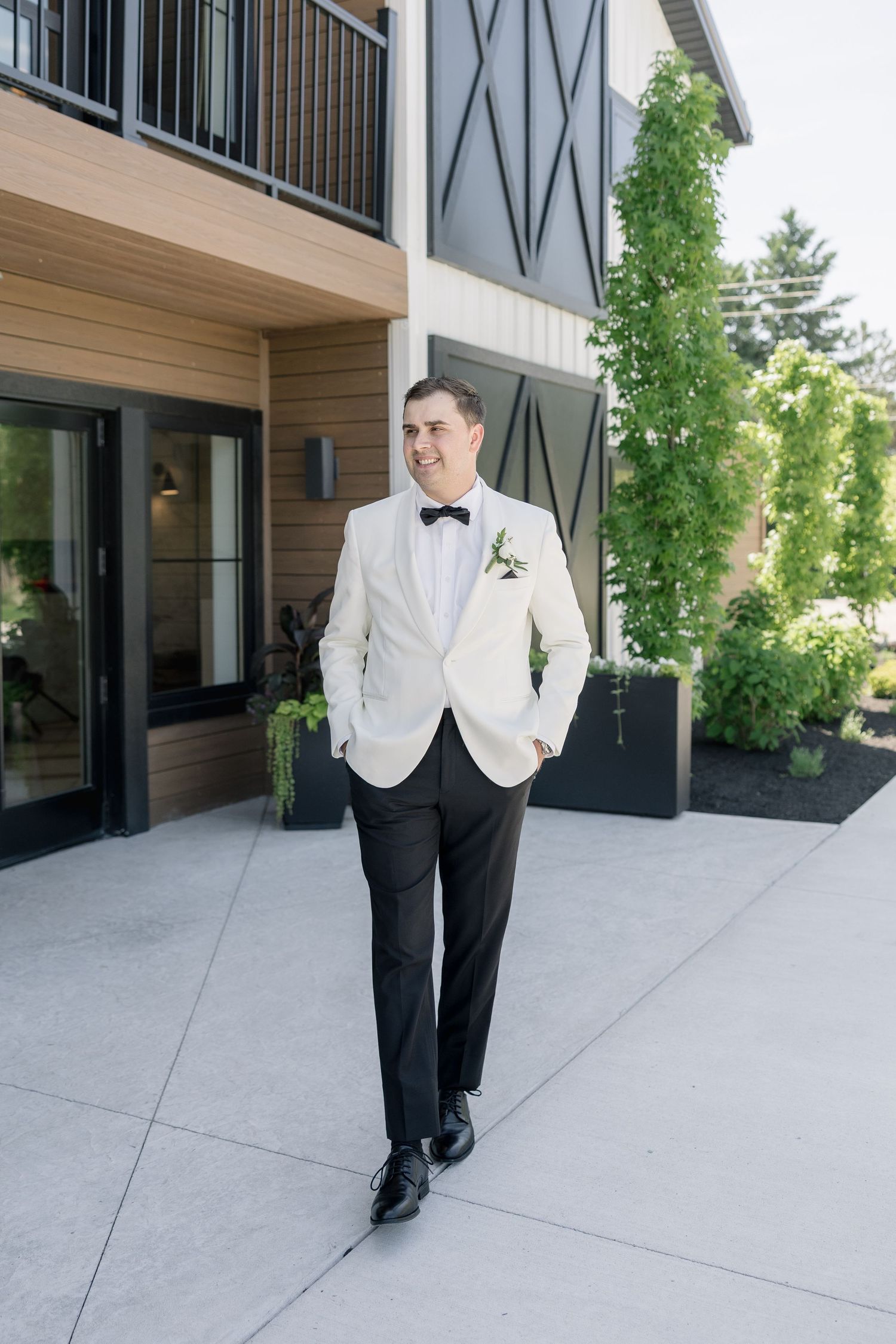 Groom in white tuxedo jacket and black pants with bow tie and boutonniere poses outdoors at modern wedding venue.