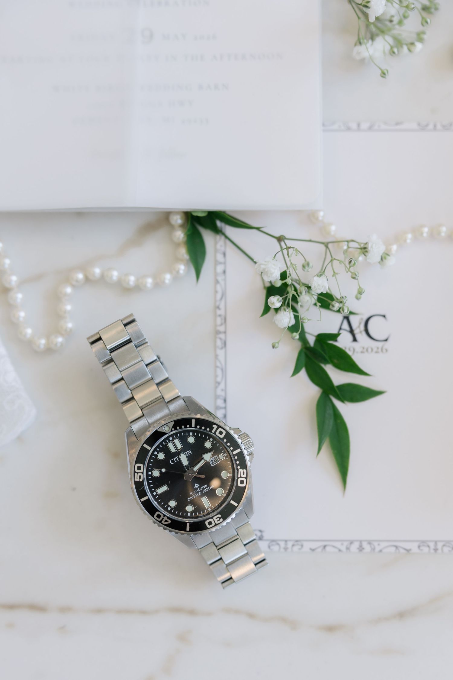 Silver diver watch on marble surface with pearl necklace, white flowers, and wedding stationery.