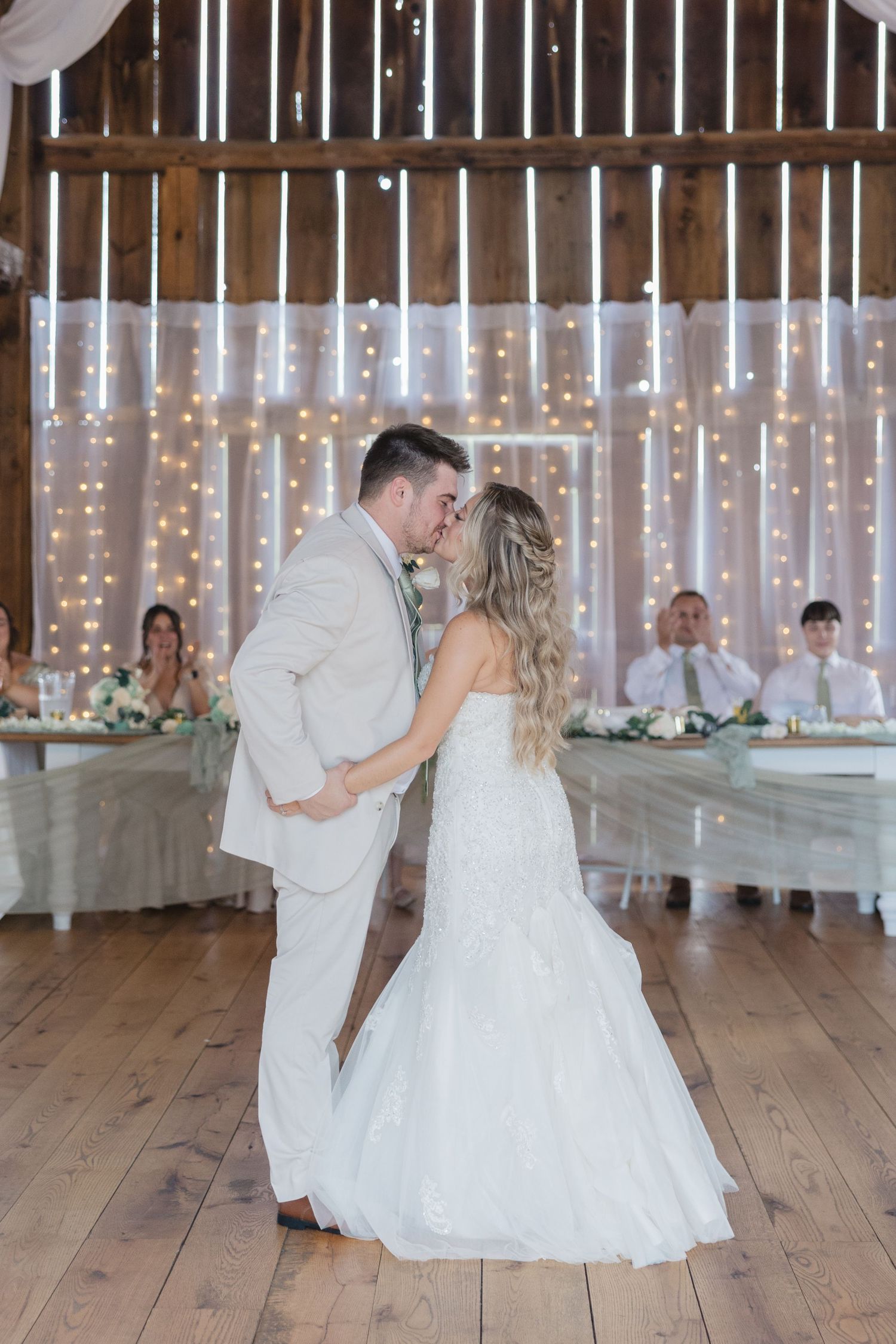 Bride and groom share first dance in rustic barn wedding venue with fairy lights backdrop.