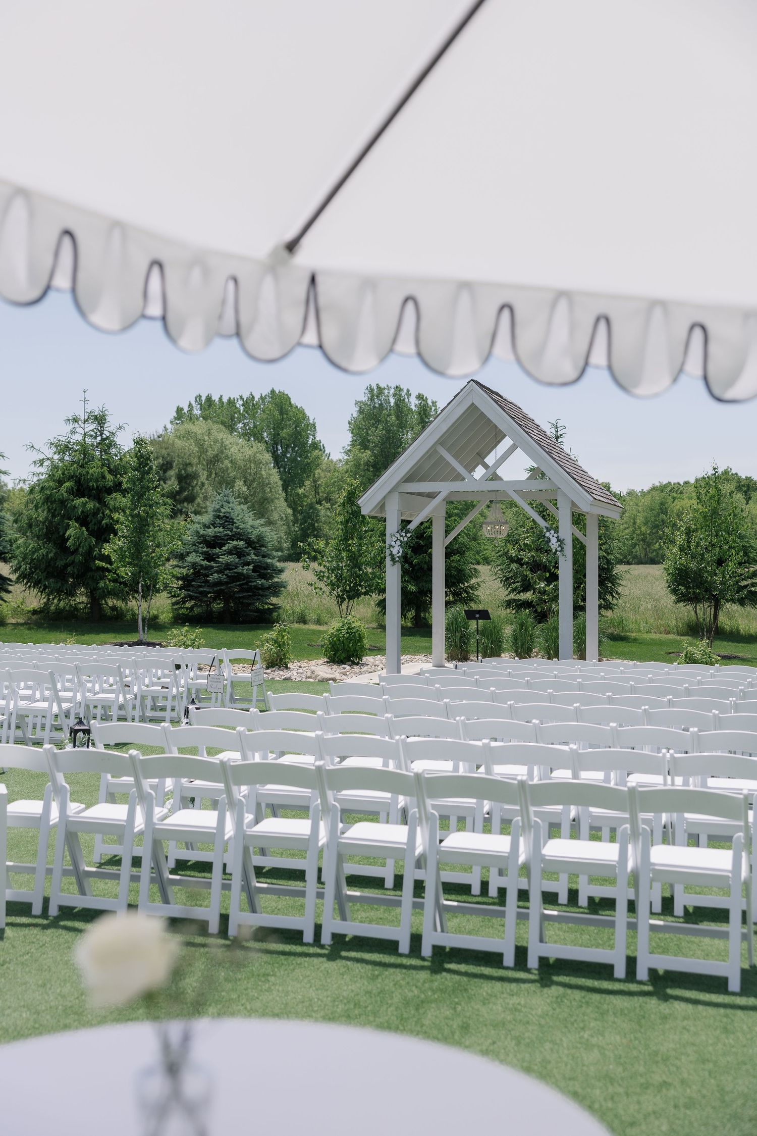 Outdoor wedding ceremony setup with white folding chairs, a wooden arbor, and lush green trees under a striped tent.