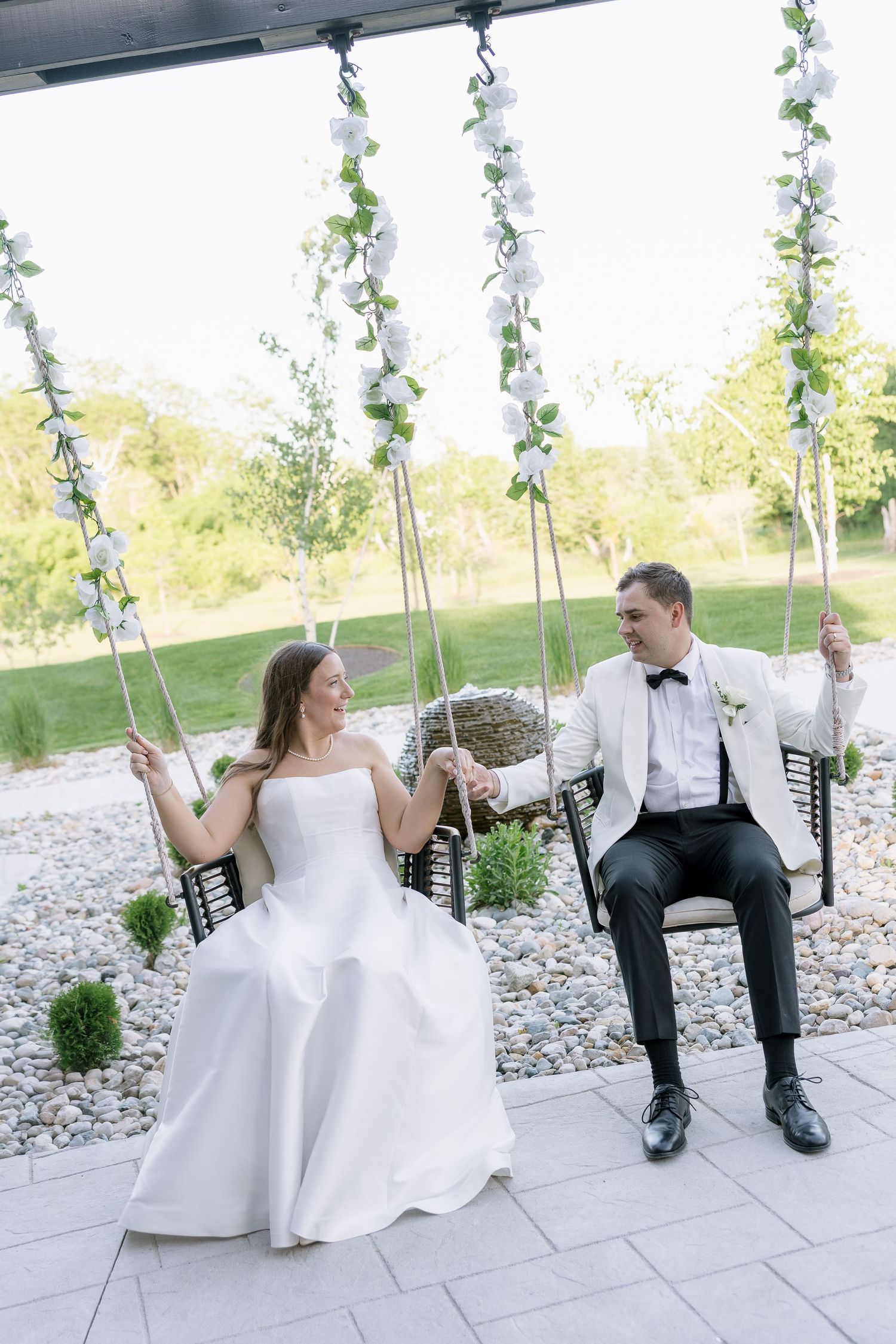 Bride and groom sit on floral swing holding hands at outdoor wedding venue with garden backdrop.