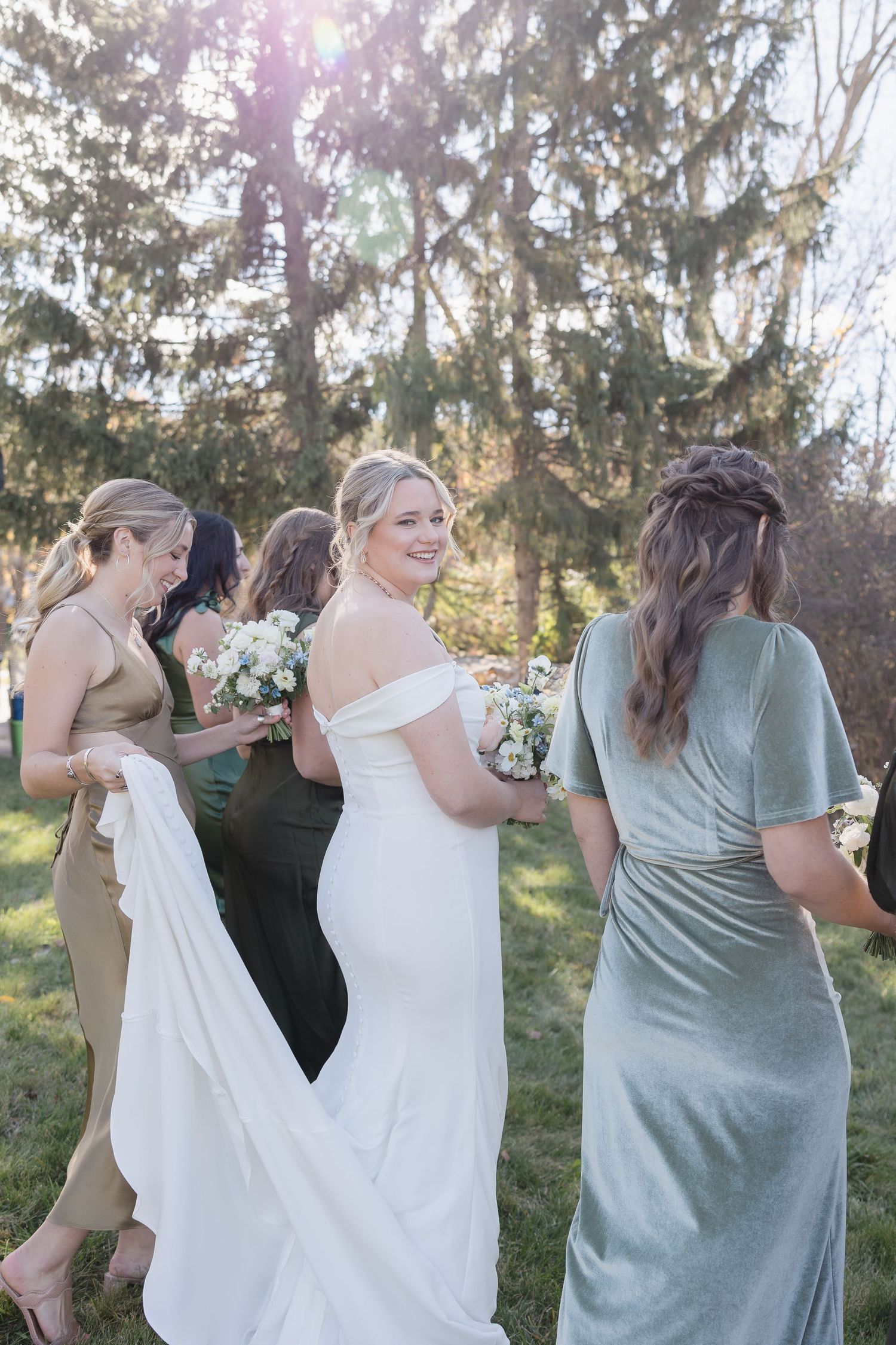 Bridal party in a mix of sage green and beige dresses pose together outdoors with bouquets on a sunny day.