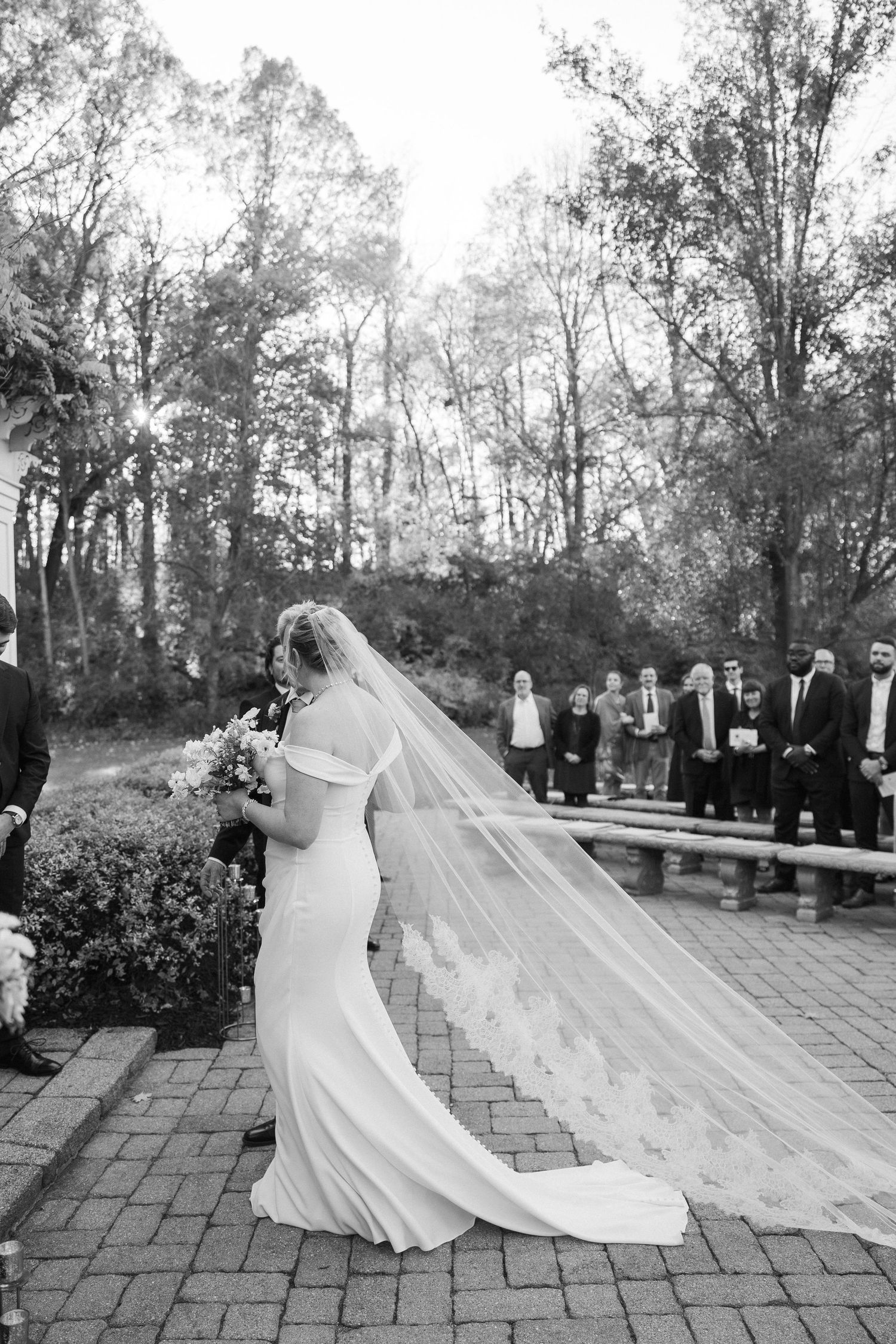 A dramatic black and white photo of a bride in a flowing dress and veil during an outdoor ceremony.