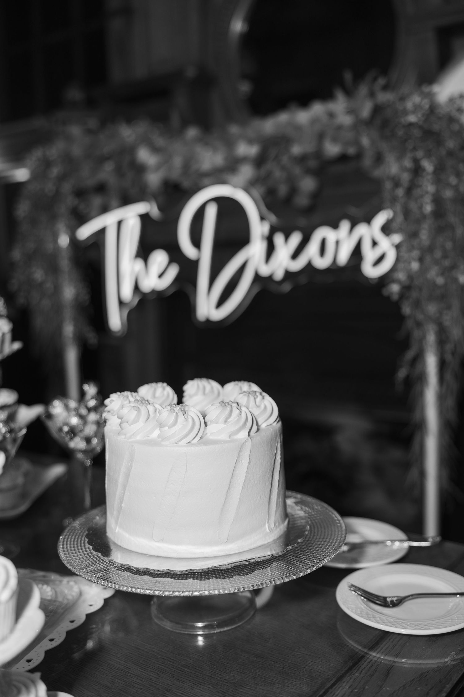 White wedding cake with piped frosting on a silver stand displayed under decorative signage reading 'The Dixons'.