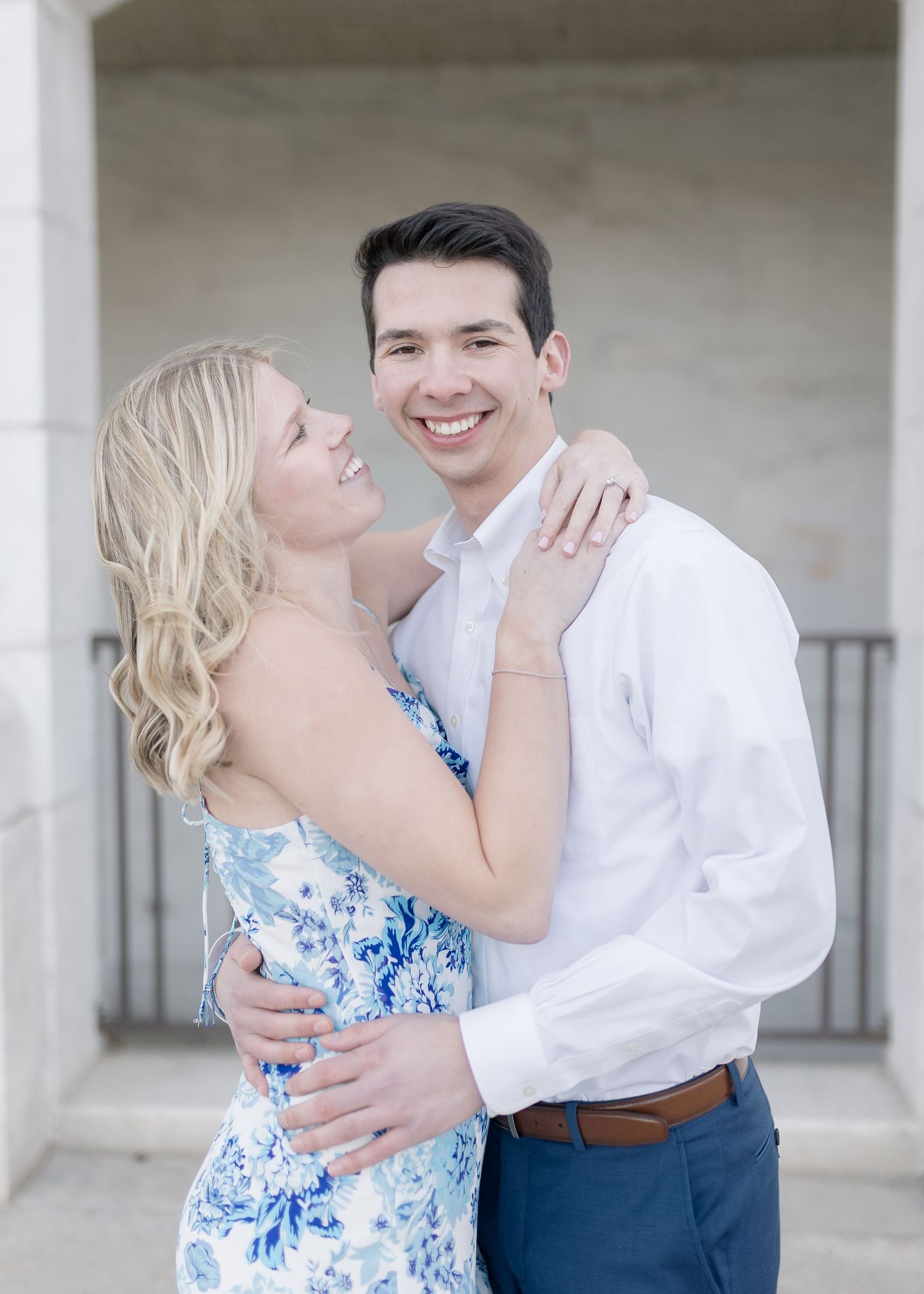 A couple embraces and smiles while posing together, with her wearing a blue floral dress and him in a white shirt.