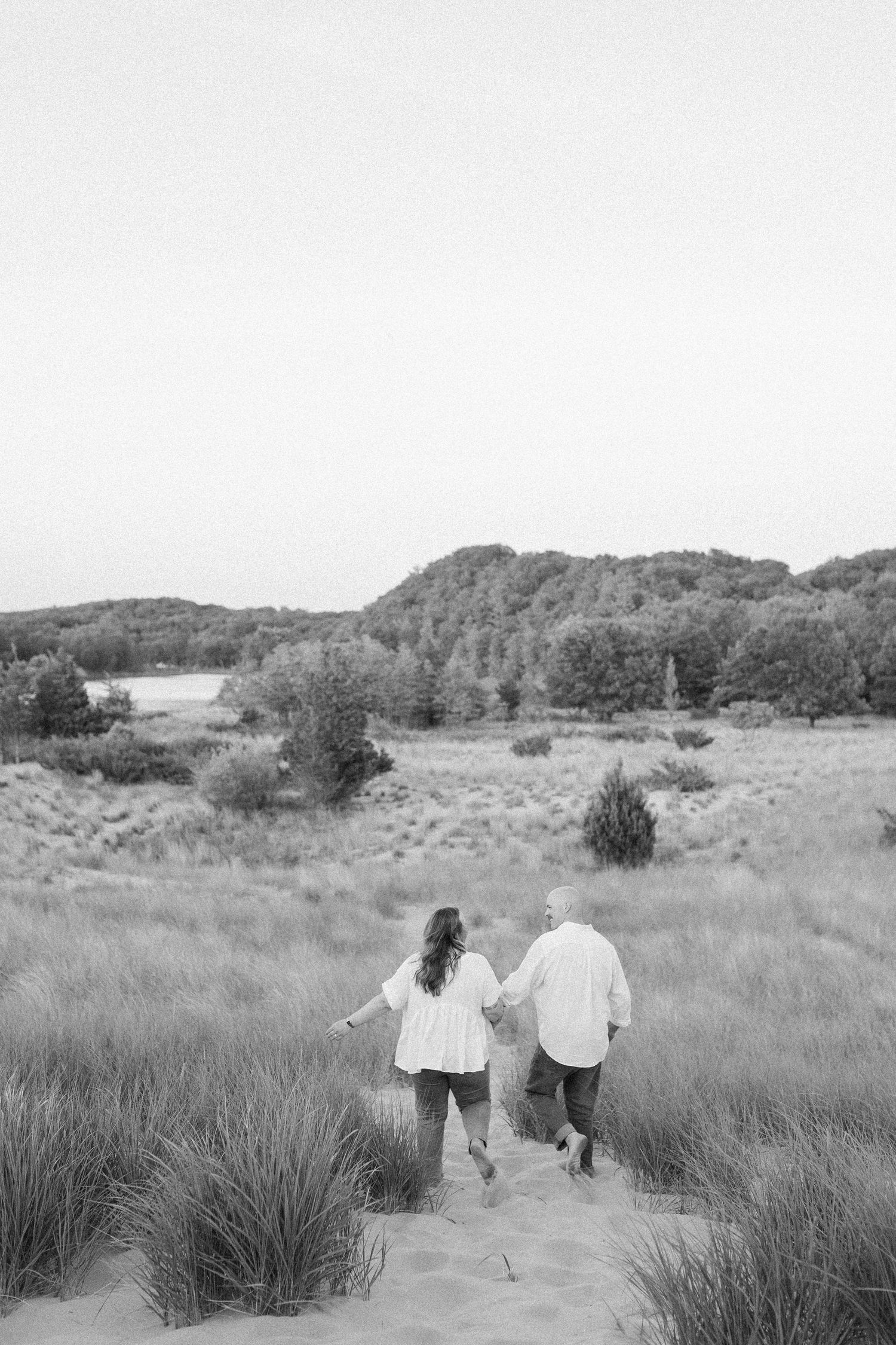 Two people walk together through a scenic grassland area with rolling hills and trees in the background.