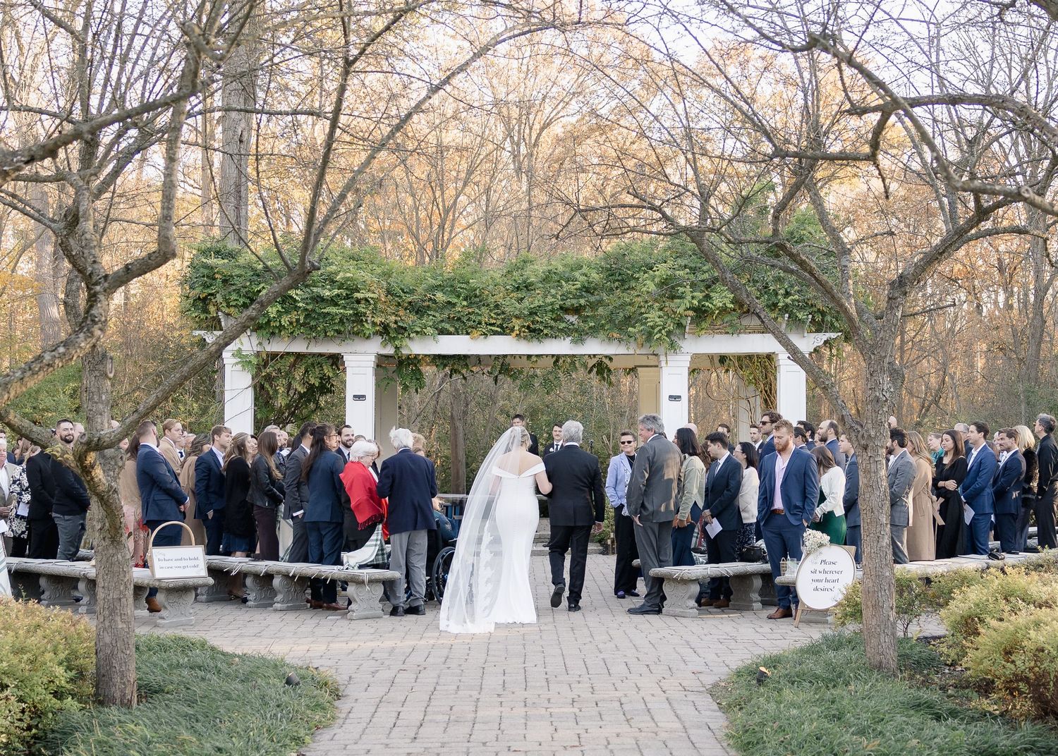 Guests gather under a white pergola decorated with greenery for an outdoor wedding ceremony.