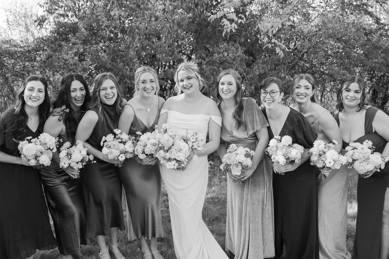 Black and white portrait of a bridal party with bridesmaids in elegant dresses holding floral bouquets.