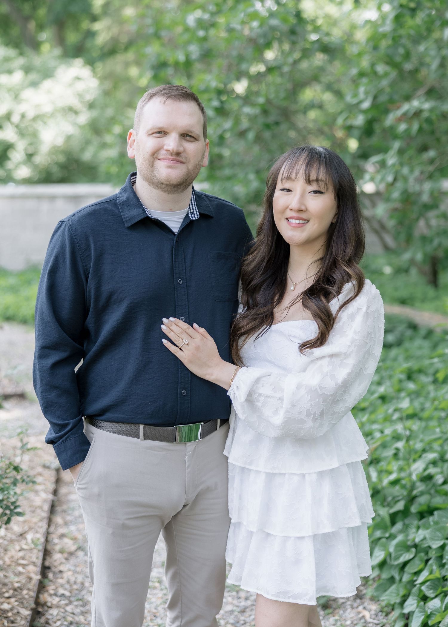 Happy couple poses together in casual attire against green garden background.