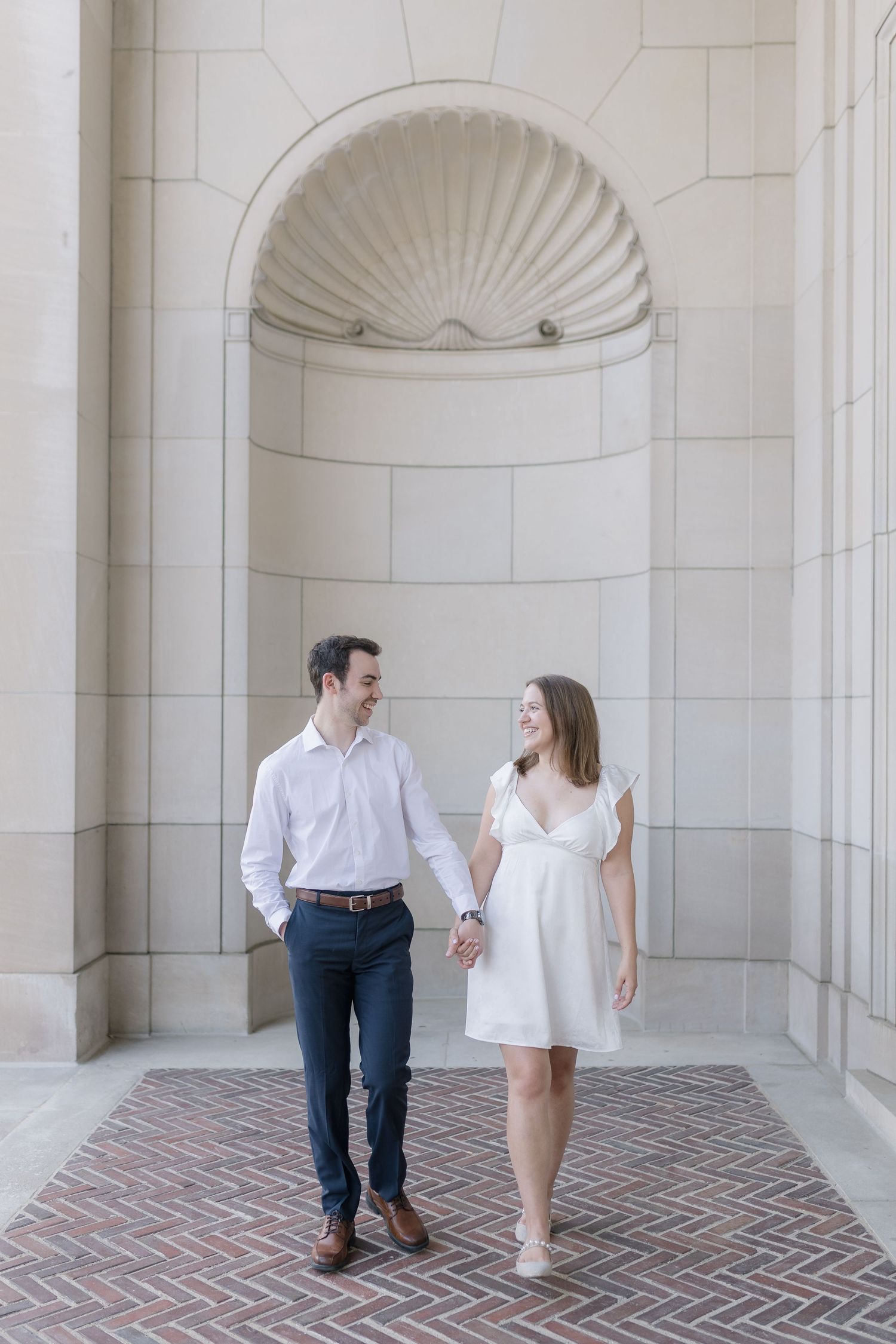 A couple shares a romantic moment in an elegant colonnade with white stone arches and brick flooring.