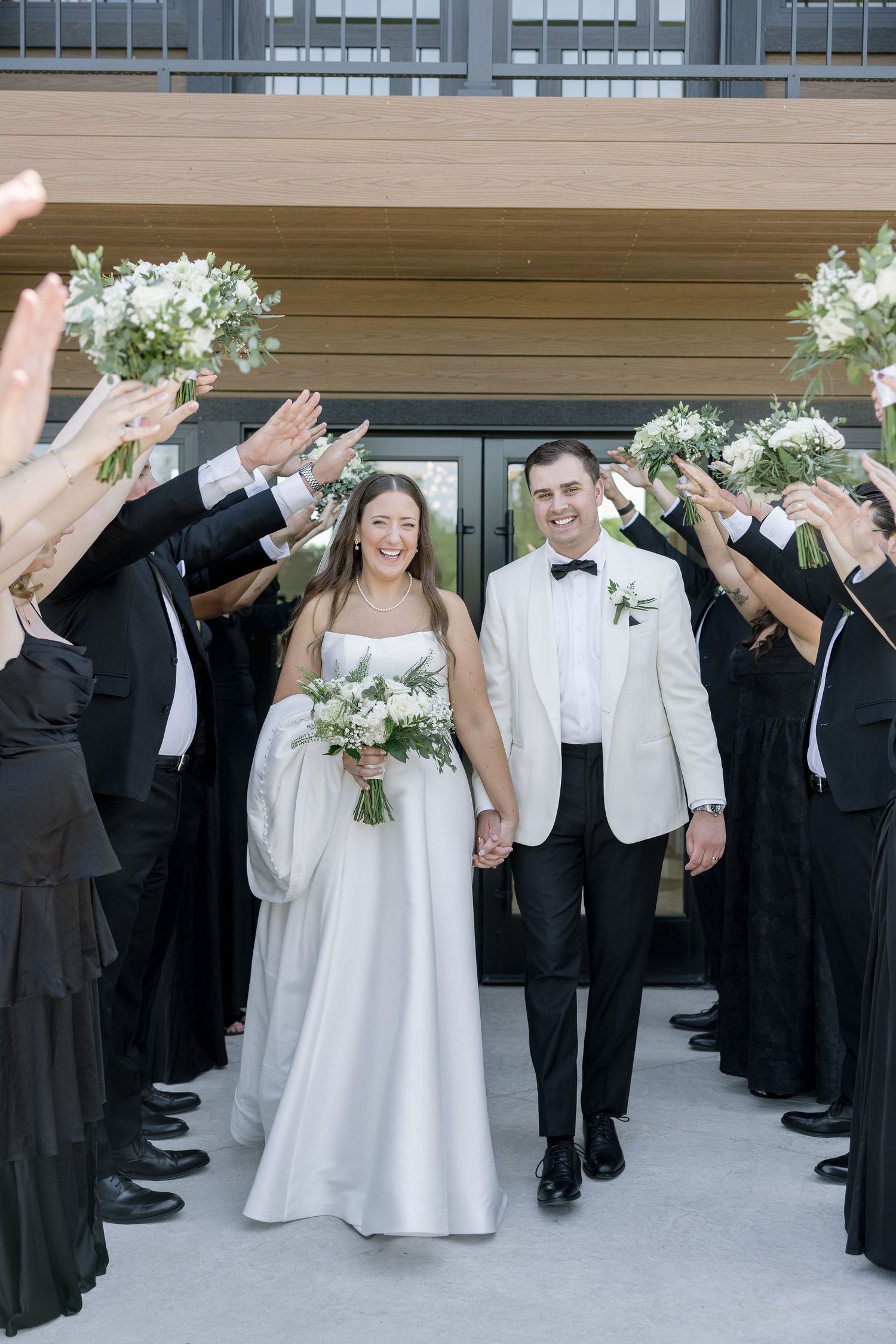 Bride and groom walk through arch of raised bouquets held by wedding party in black and white formal attire.