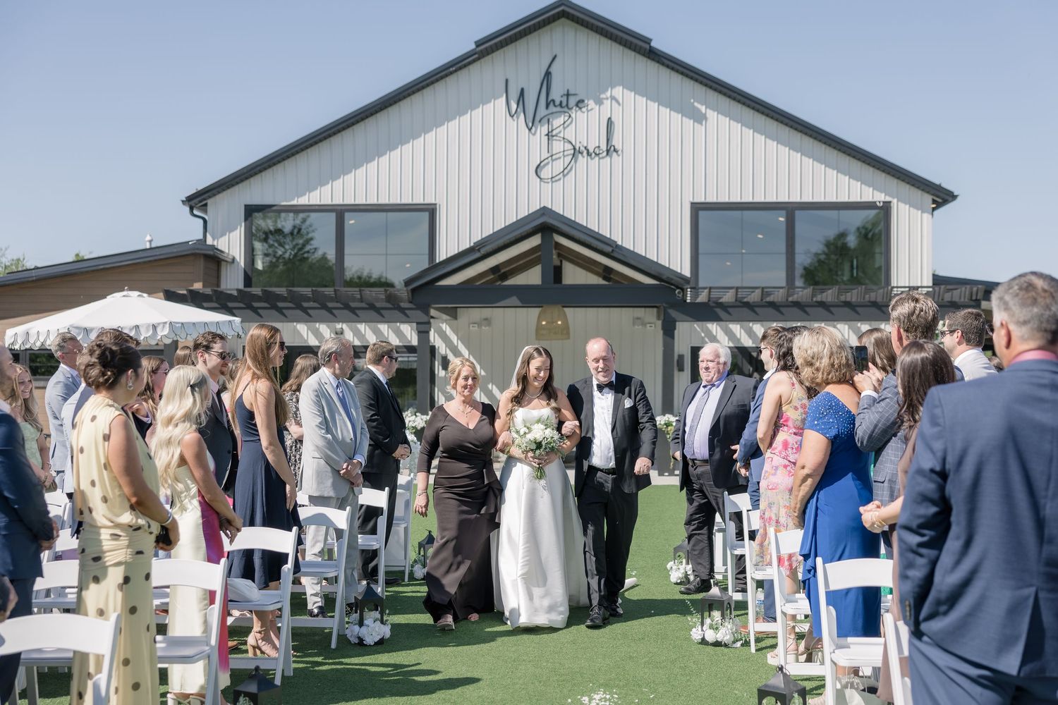 Bride and groom walk back down the aisle at White Barn wedding venue as guests cheer in sunny outdoor ceremony.