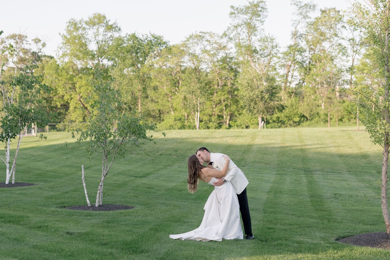 Bride and groom share a romantic dip kiss on a lush green lawn surrounded by trees on their wedding day.