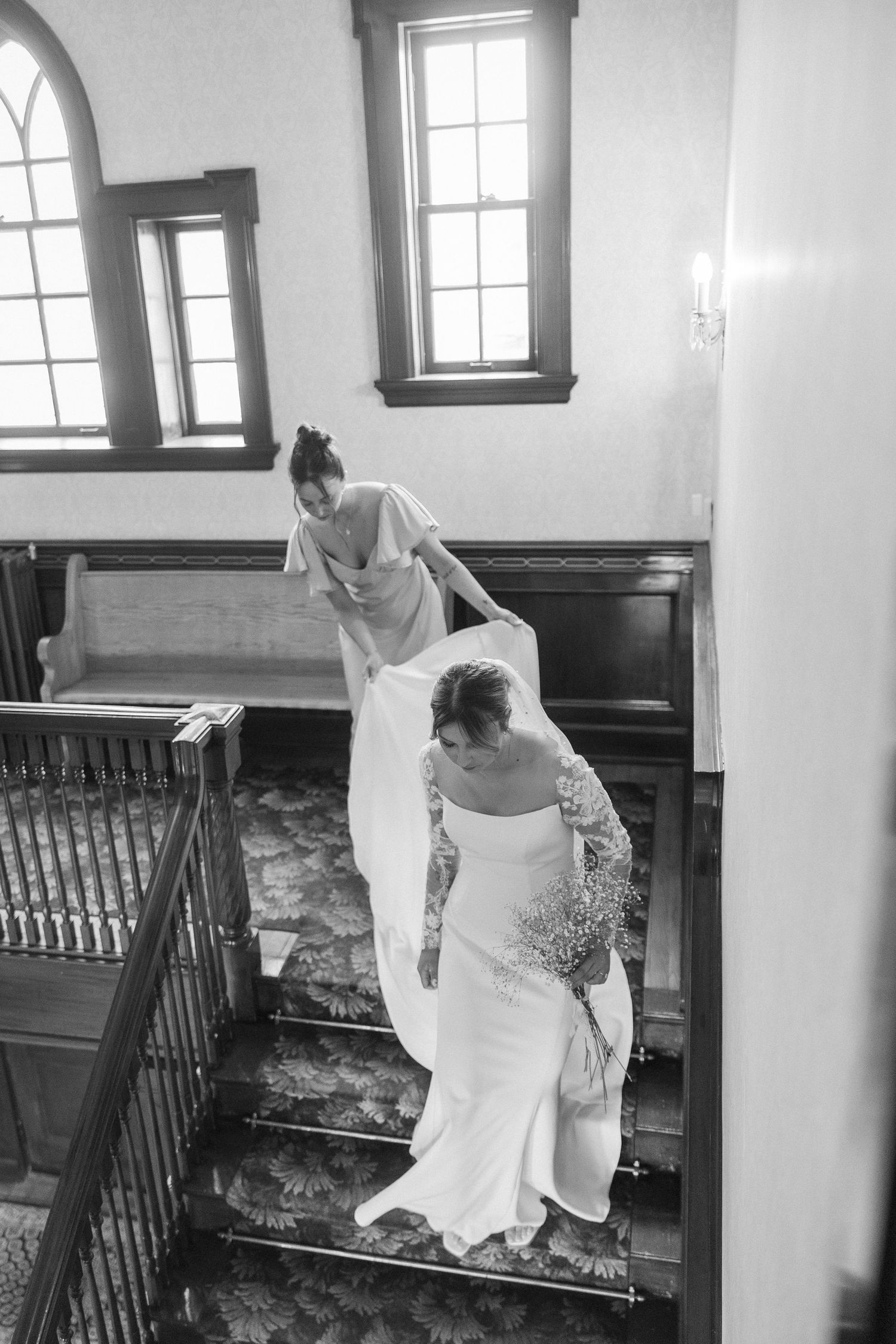 Two people in wedding attire carefully descending a grand staircase in black and white.