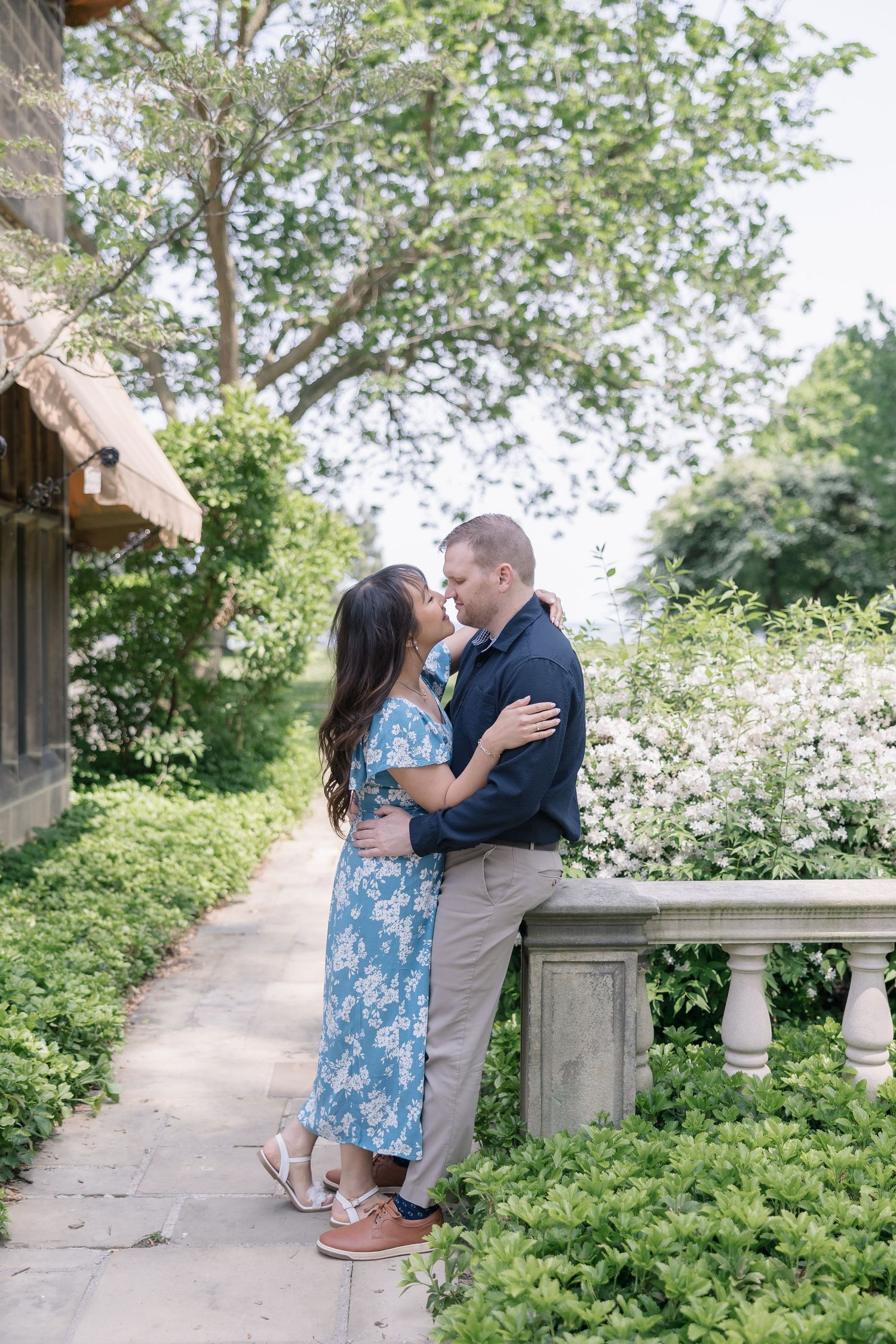 Couple shares romantic moments on stone path surrounded by lush greenery and white flowering trees in springtime.