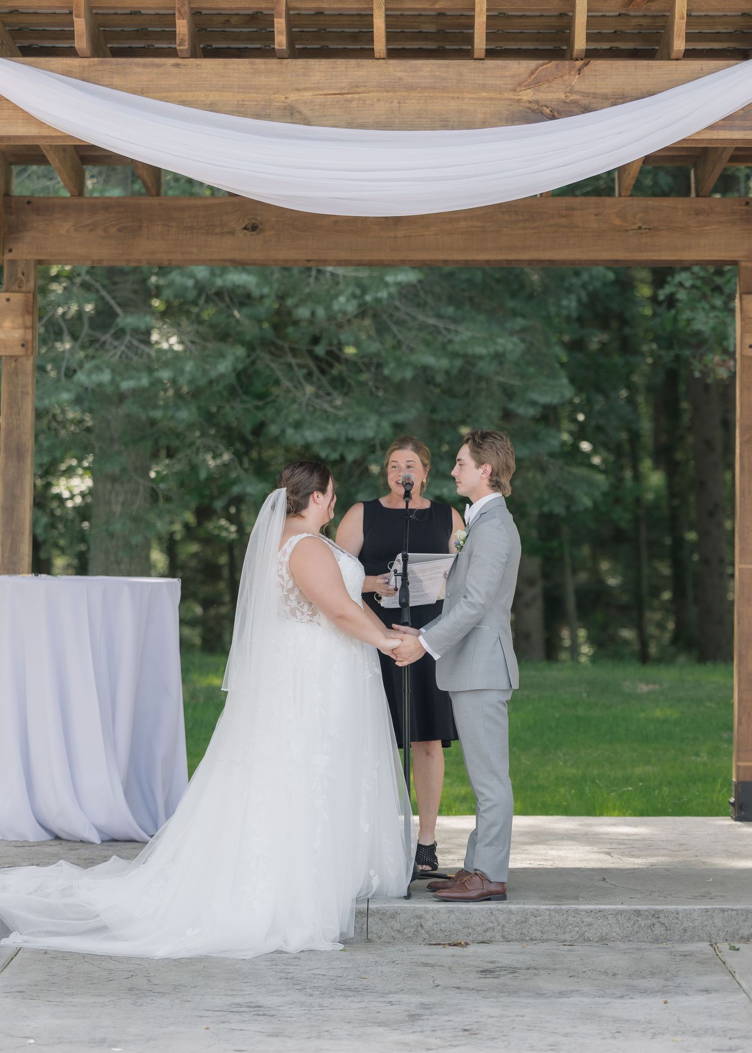 A wedding ceremony takes place under a white fabric draped wooden arch in an outdoor garden setting.