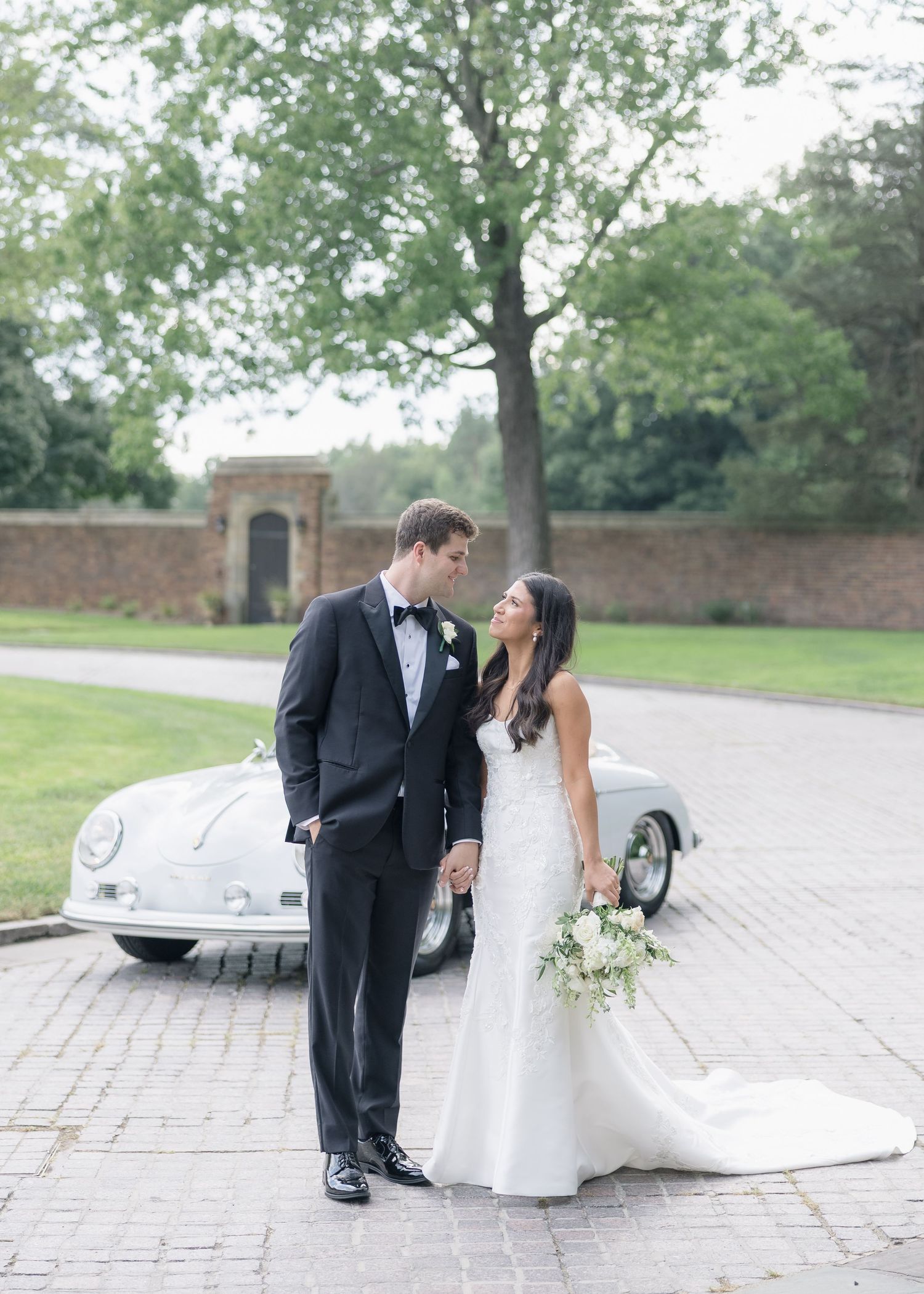 A romantic wedding couple poses beside a classic white vintage car on a brick paved driveway near a garden wall.