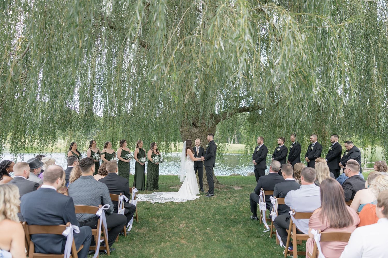 An outdoor wedding ceremony takes place under a graceful weeping willow tree with guests seated on white chairs.