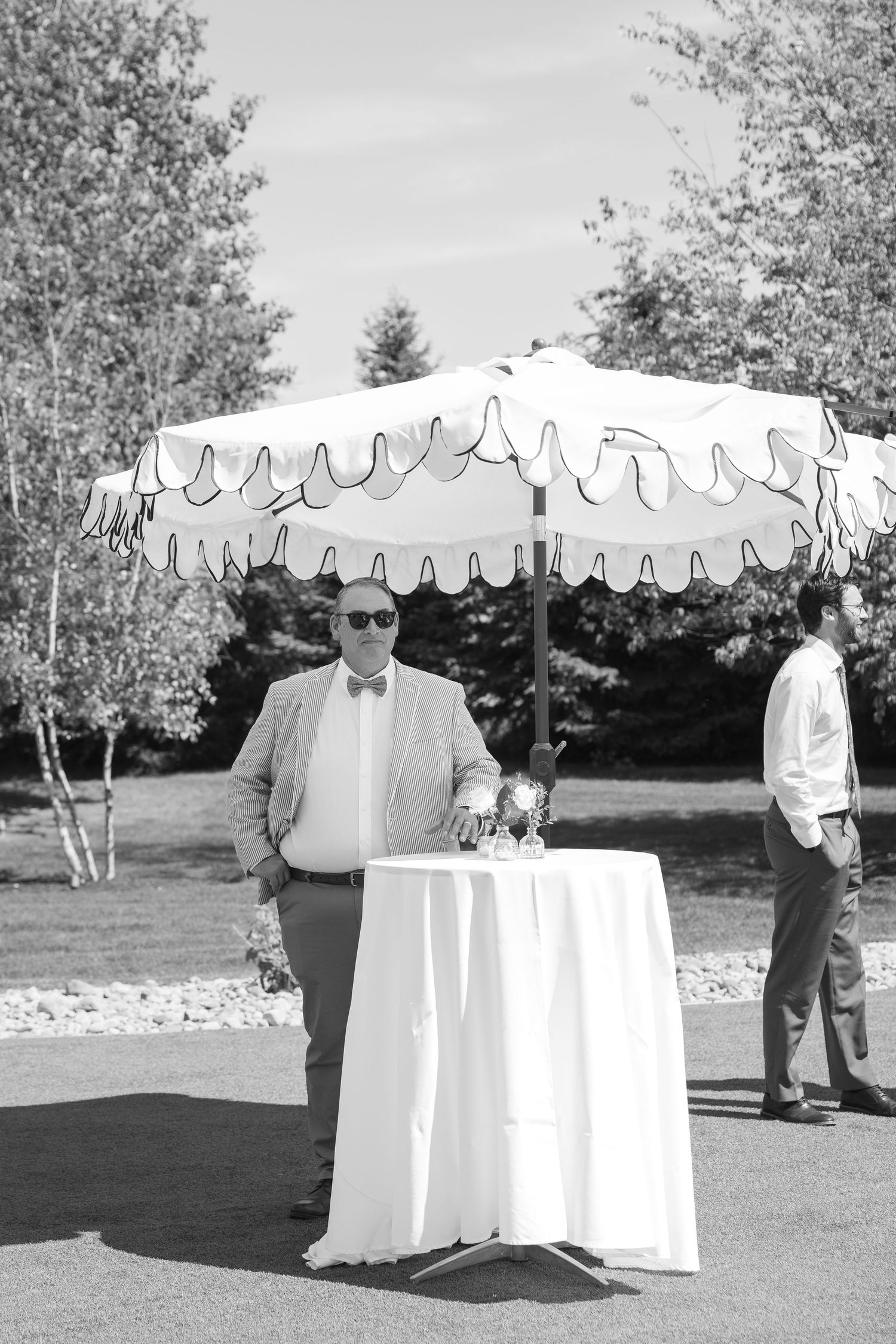 Man in bow tie and sunglasses stands by cocktail table under decorative umbrella at outdoor wedding reception.