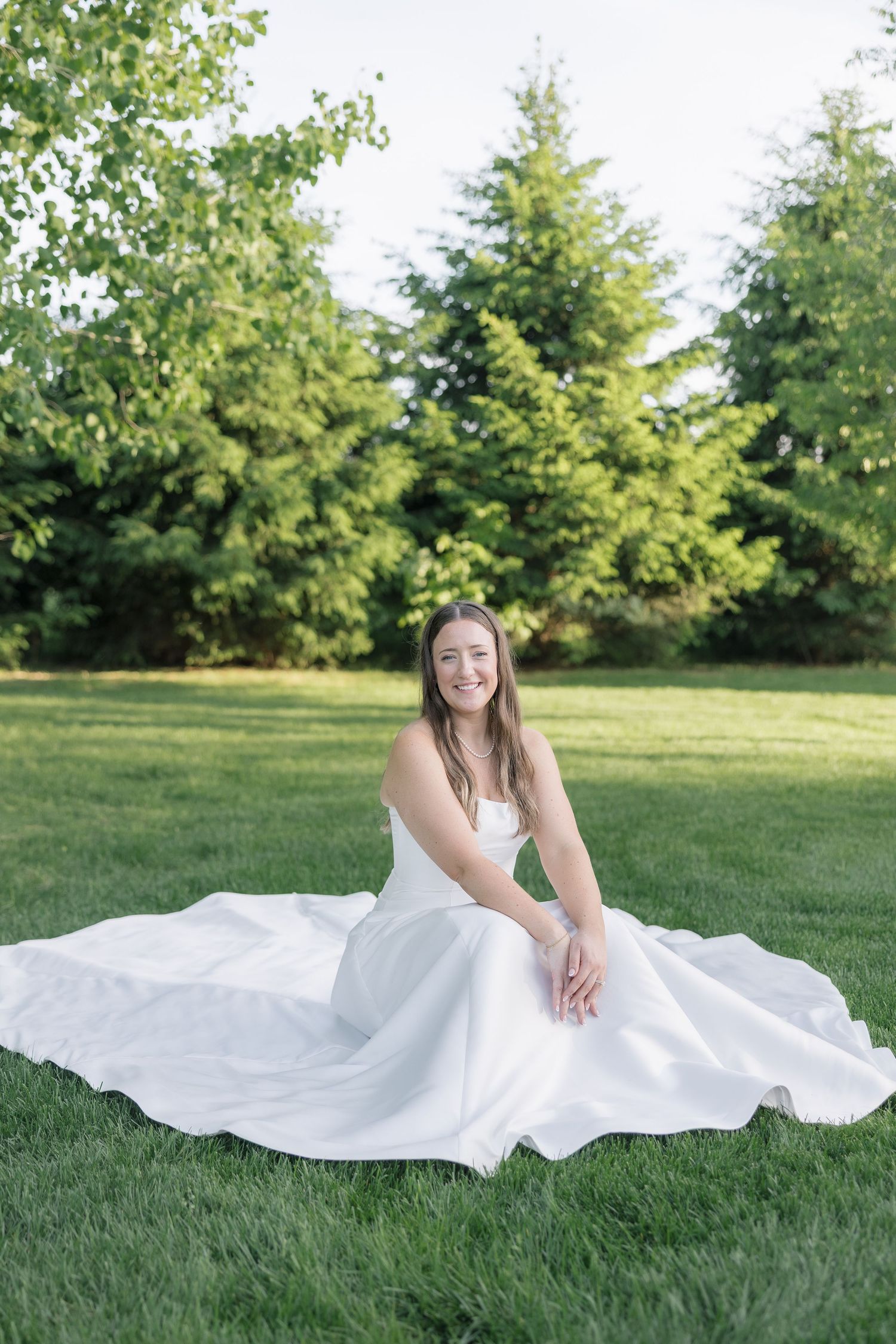Smiling bride in white gown sitting on green grass outdoors with lush trees in background on a sunny day.