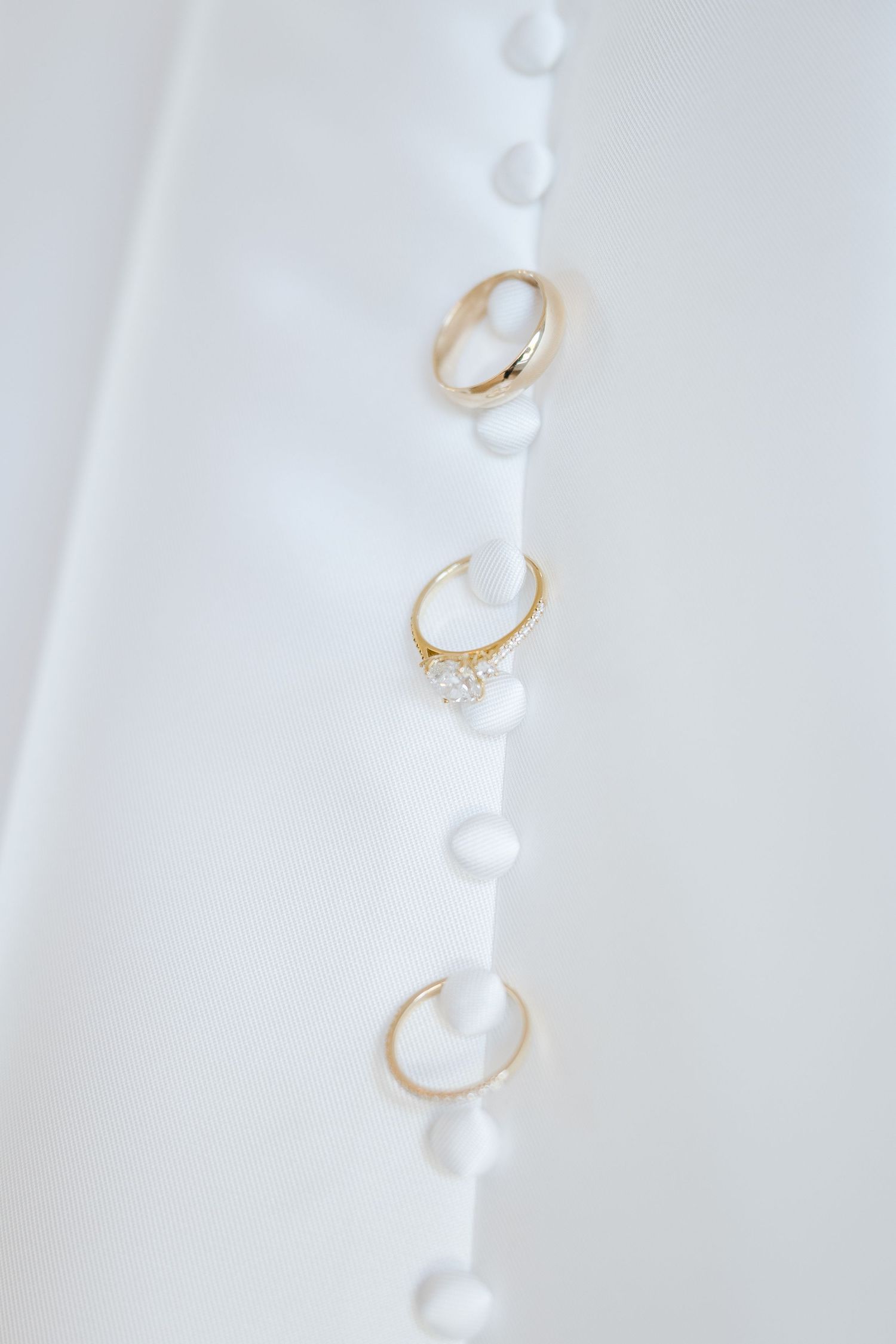 Three gold wedding rings displayed vertically on white fabric with pearl button detail.