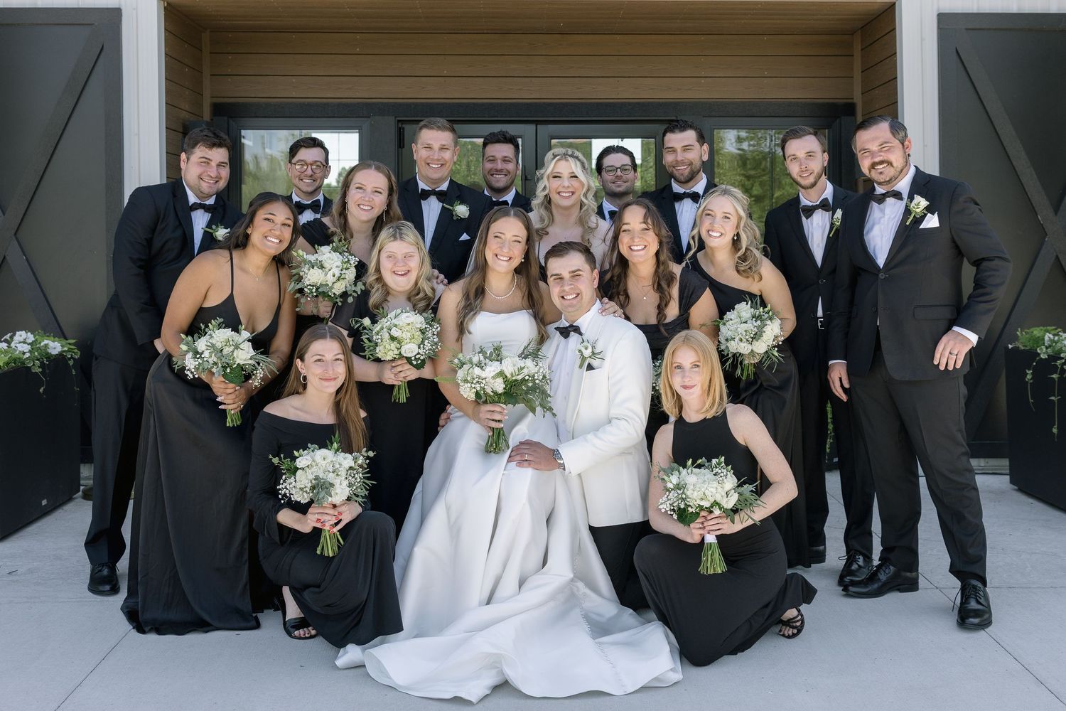 Wedding party in black and white attire posing together, bride in white gown holding bouquet of white flowers.