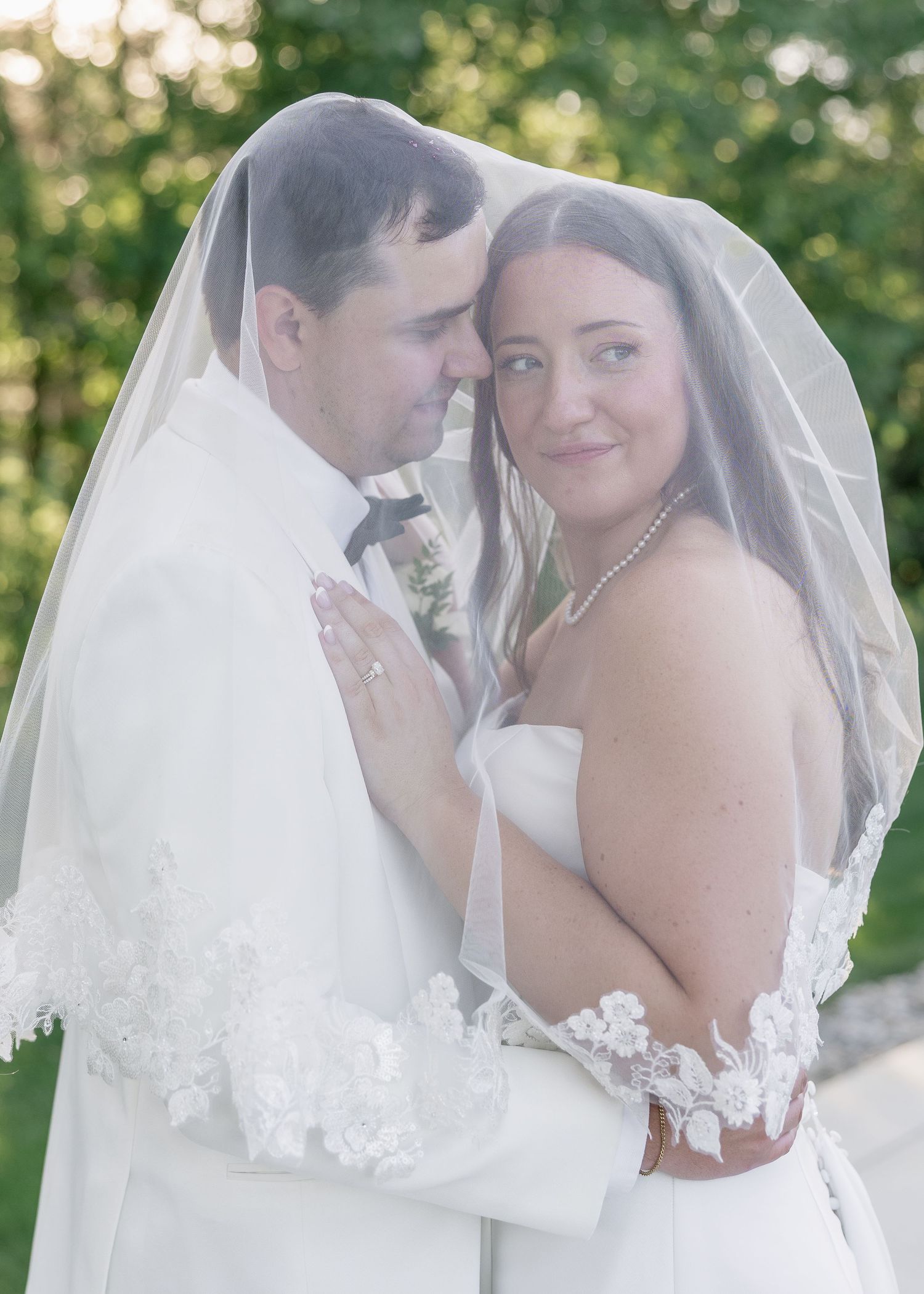 Romantic wedding portrait of bride and groom beneath lace-trimmed veil, embracing in soft golden light.