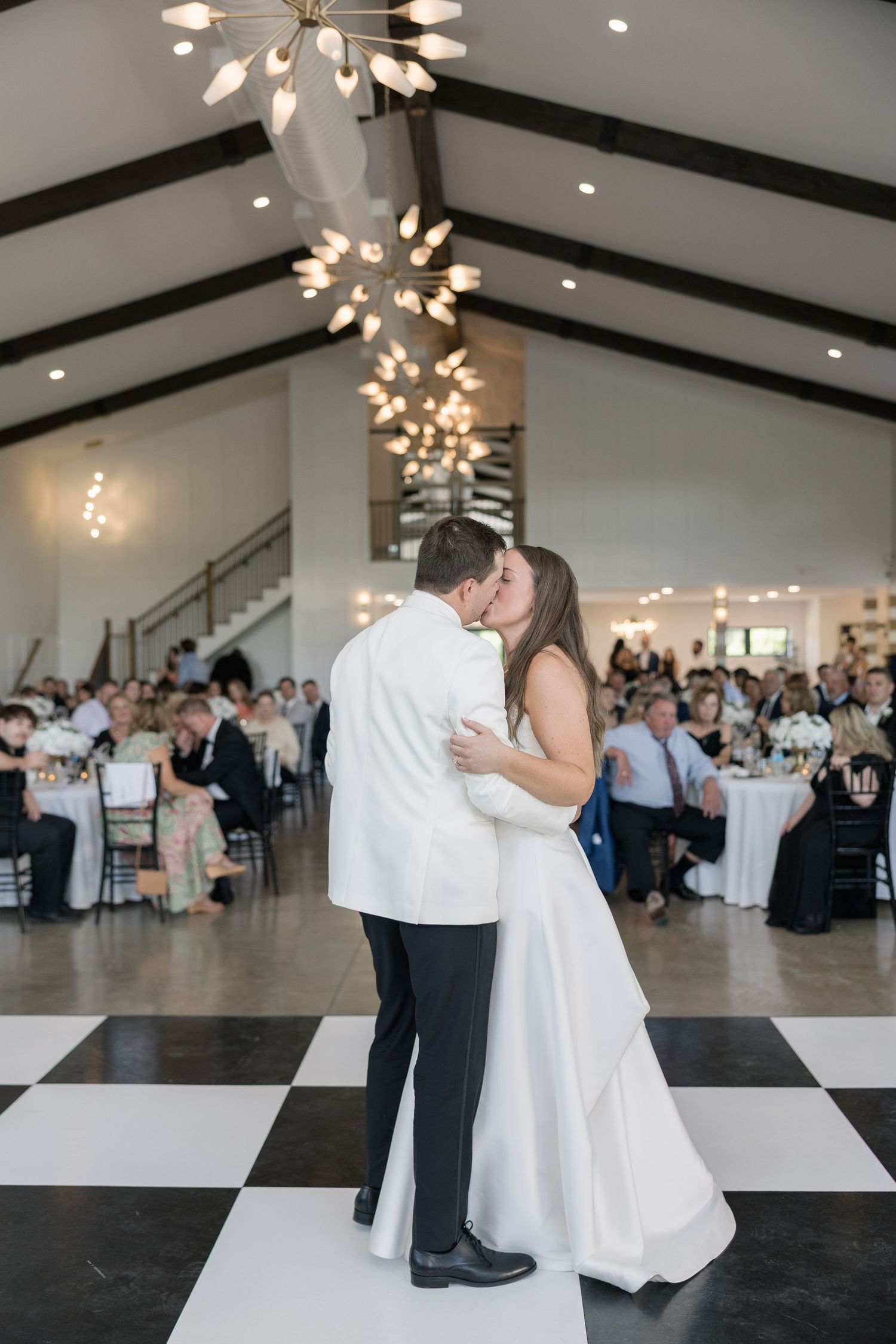 Bride and groom share first dance kiss on checkered floor in elegant white wedding reception venue with chandelier.