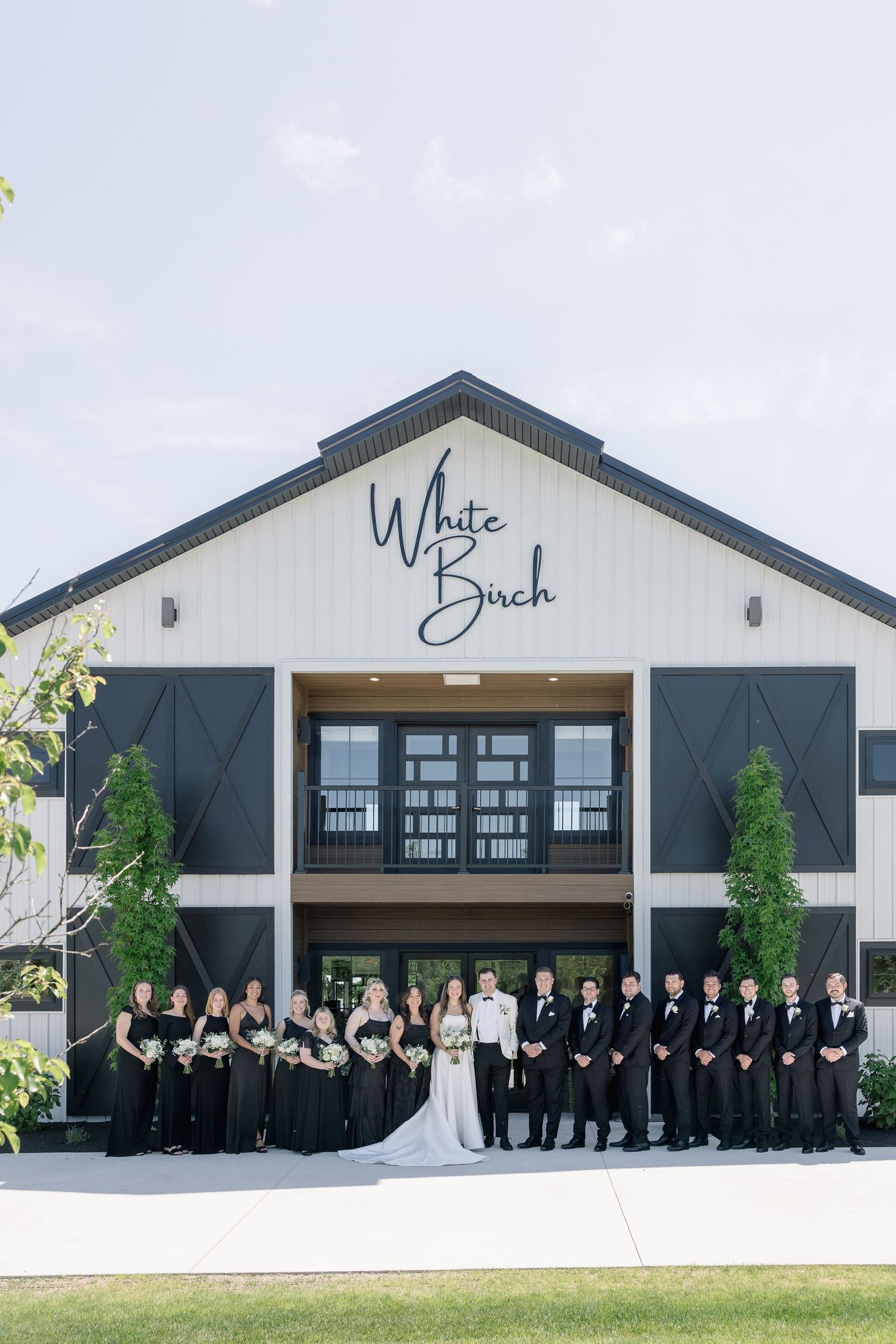 Wedding party in black attire poses outside White Birch venue on sunny day.