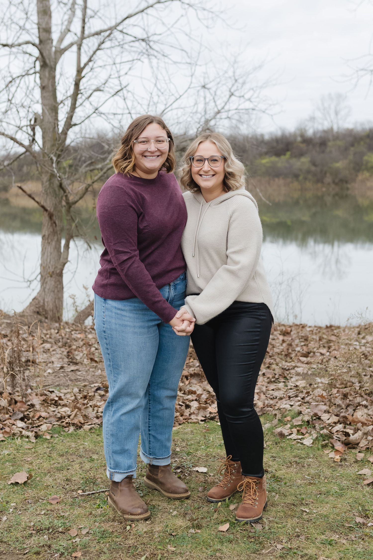 Two people in coordinating burgundy and beige outfits stand together on fallen autumn leaves by a lake.