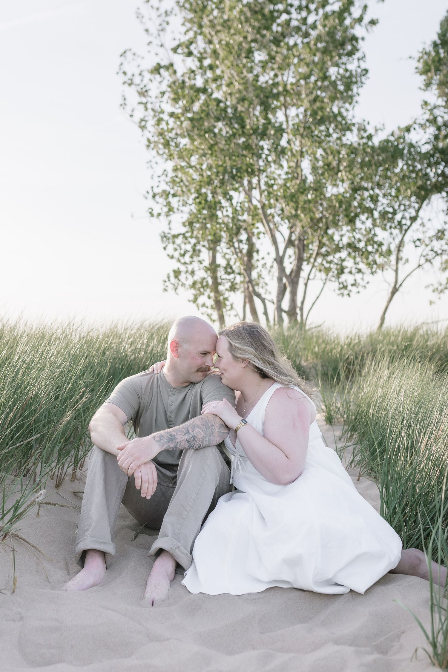 A couple sits intimately together on sand dunes with tall grass and birch trees in the background at sunset.
