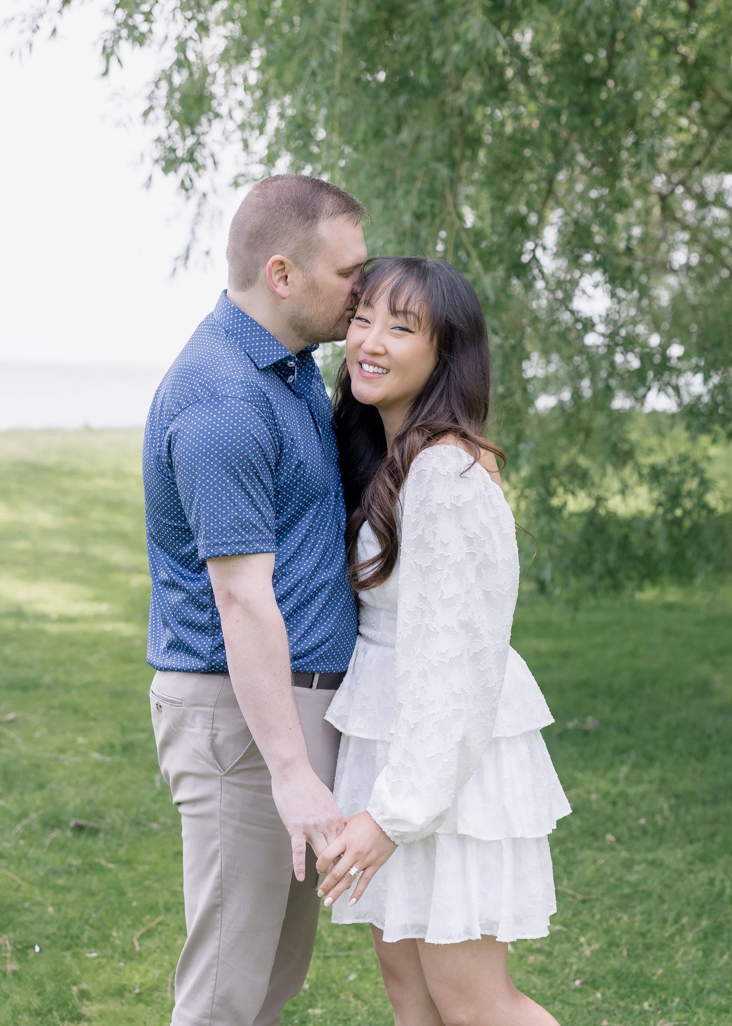 A romantic couple shares an intimate moment under willow trees in a sunlit garden setting.