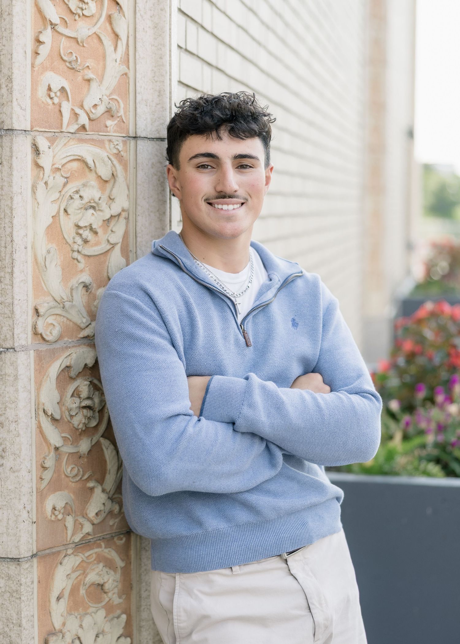 Young person in light blue sweater stands against ornate architectural columns with floral decorations in background.
