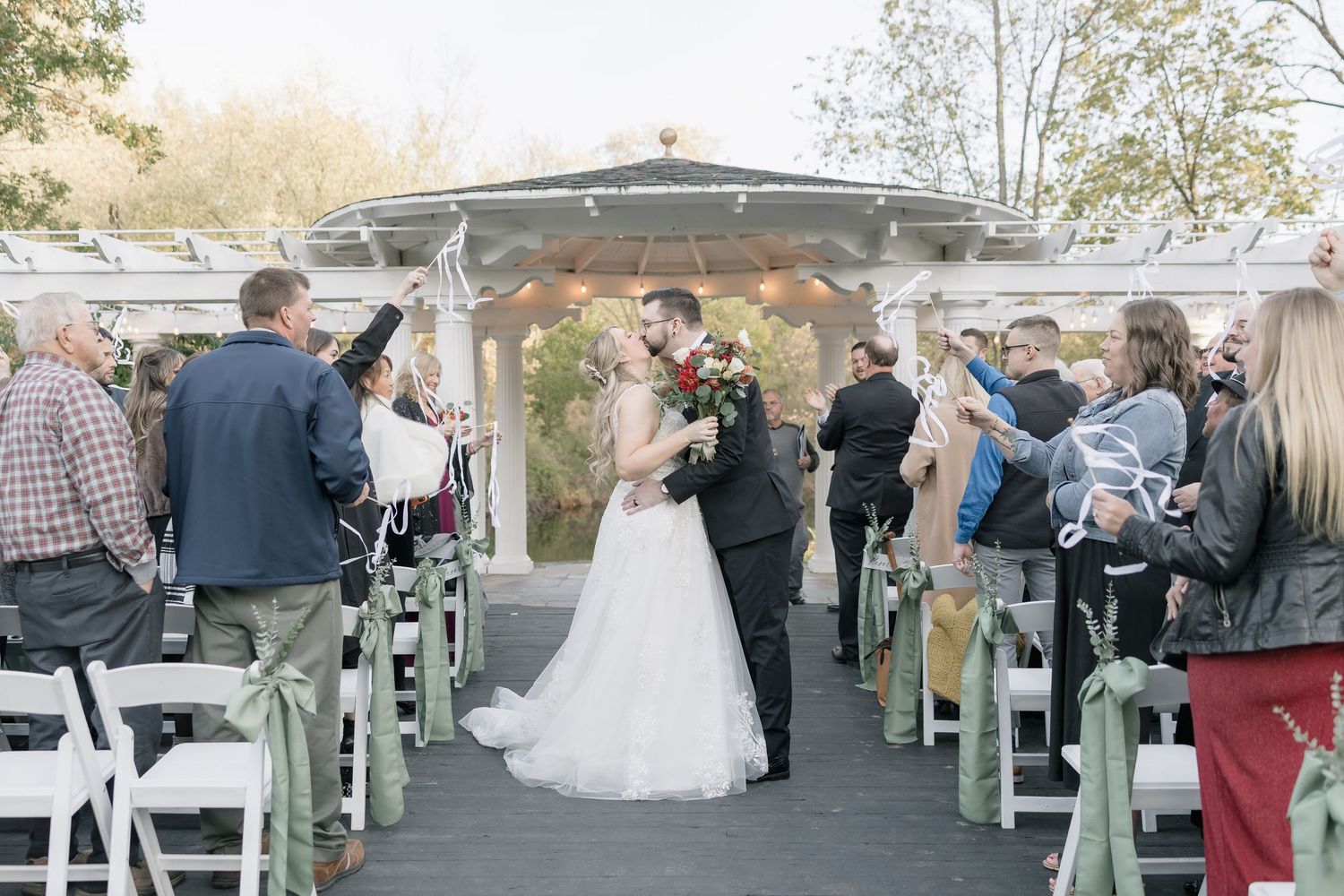 An outdoor wedding ceremony takes place under a gazebo with guests seated in white chairs on a wooden deck.