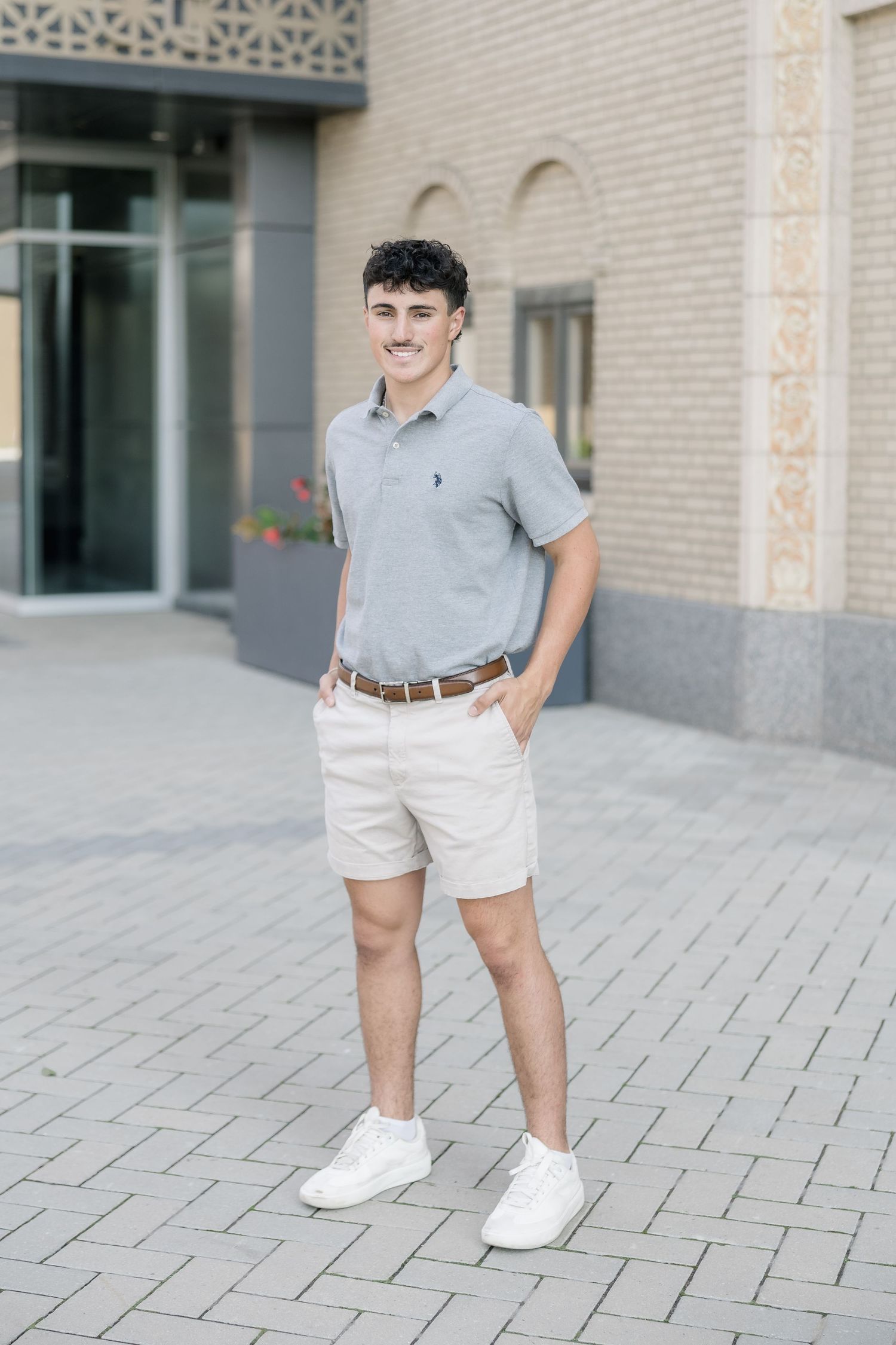 A person wearing a light gray polo shirt and white shorts stands on a brick paved walkway outside a building.