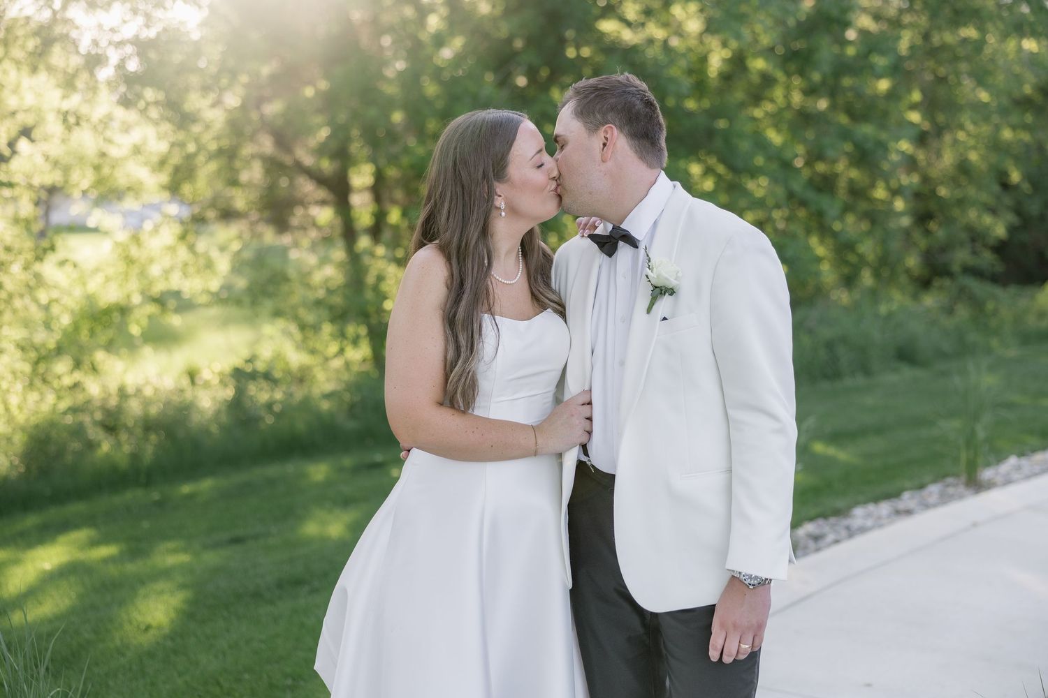 Bride and groom sharing a romantic kiss on a sunlit pathway surrounded by beautiful green trees.