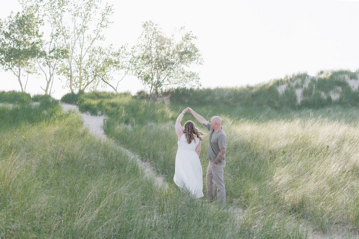 A couple in wedding attire dances together in a dreamy field of tall grass at sunset.