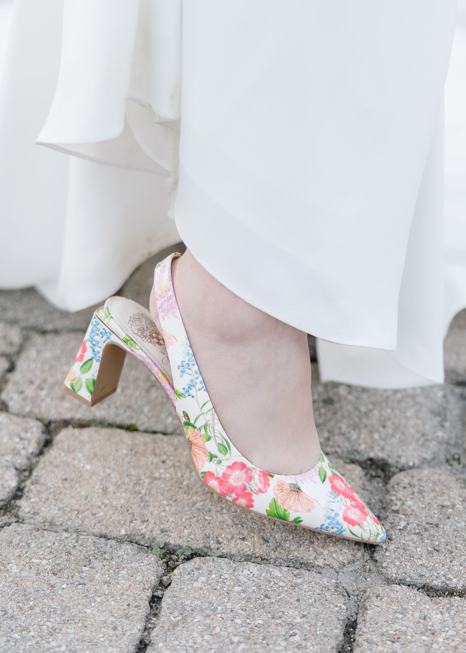 Floral patterned wedding shoes with a low heel on stone pavement beneath a white dress.