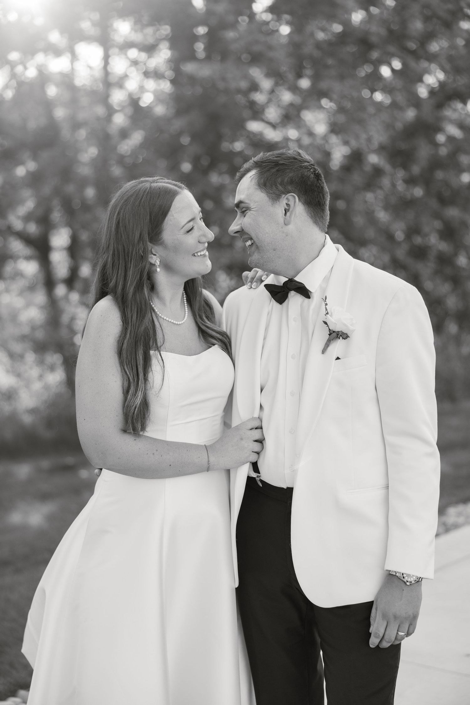 Black and white photo of a smiling bride and groom in white jacket and bow tie gazing at each other outdoors.