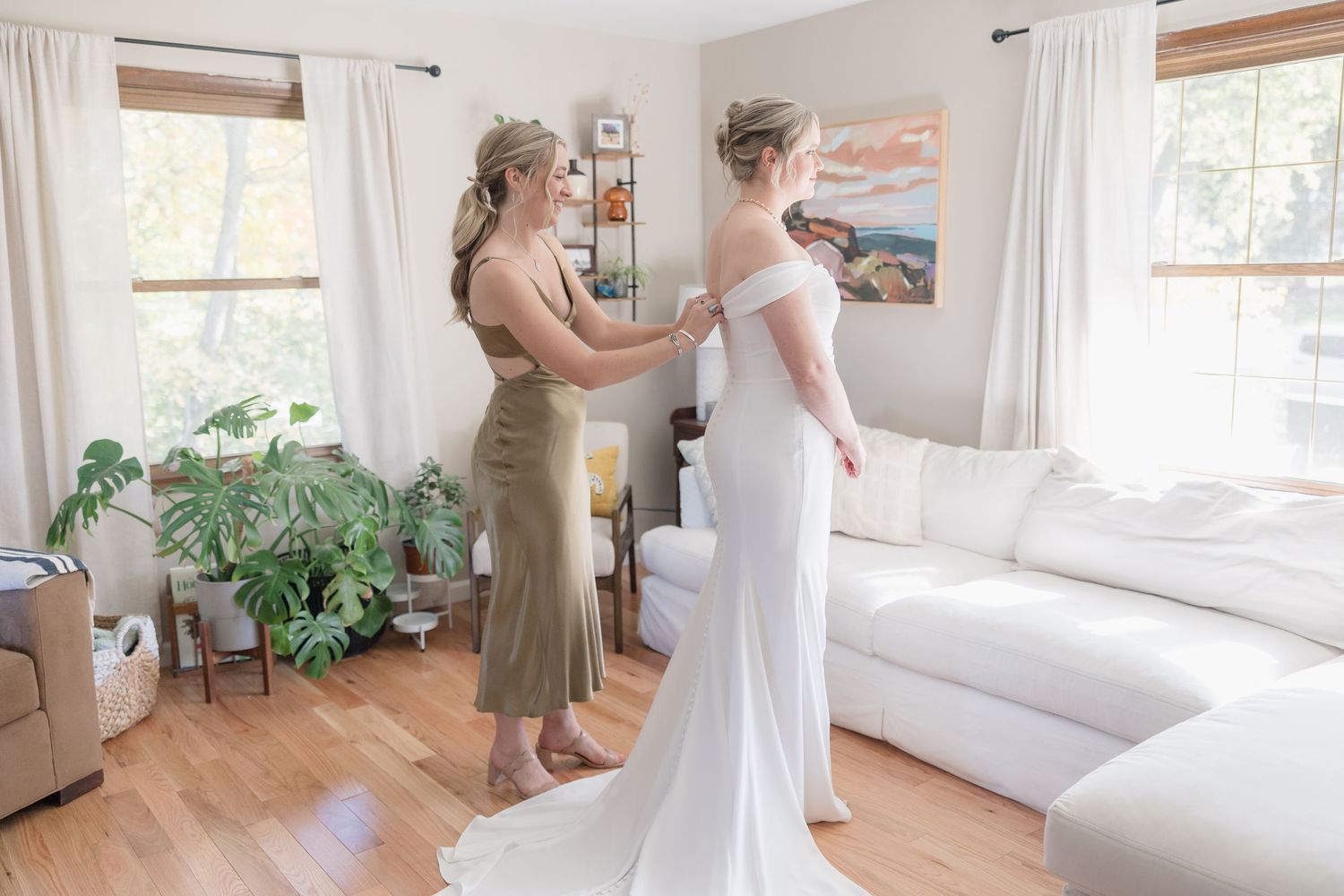 A formal dress fitting scene in a bright, modern living room with houseplants and white decor.