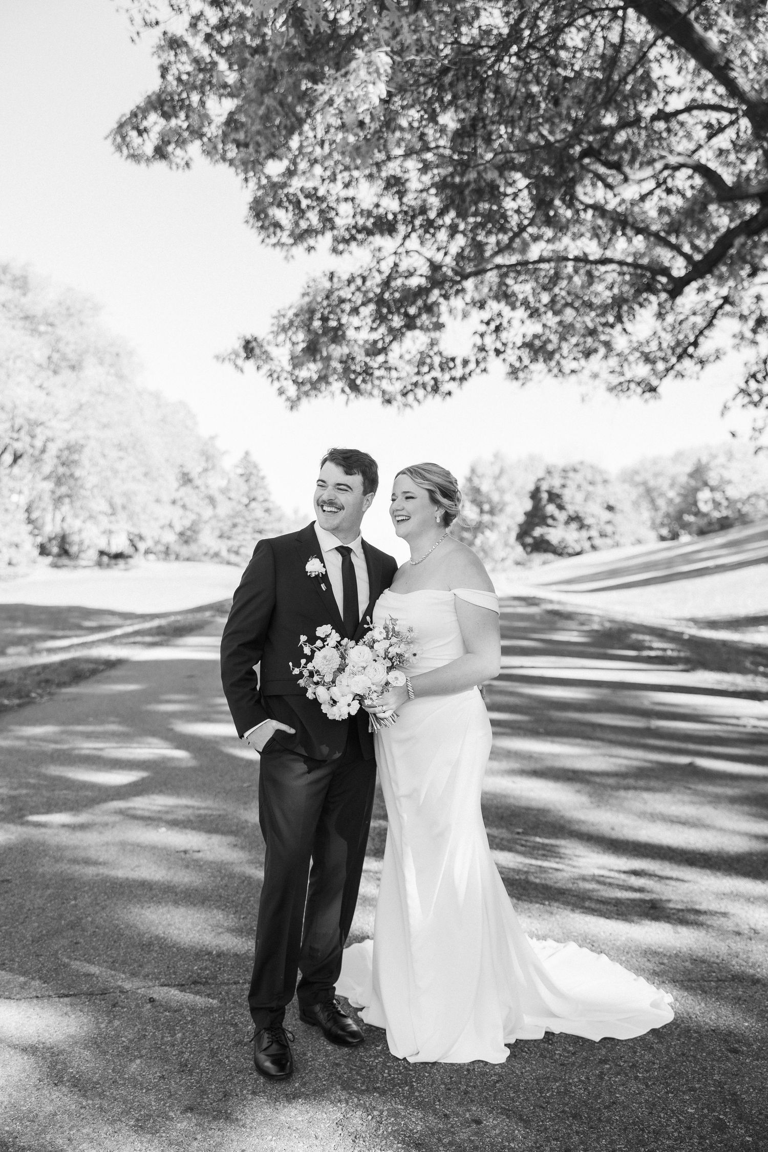 Black and white portrait of newlyweds standing together on sunlit wedding day path.