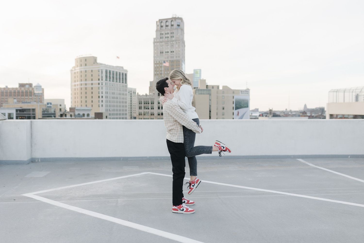 A playful lifting pose on a rooftop parking area with city buildings behind.