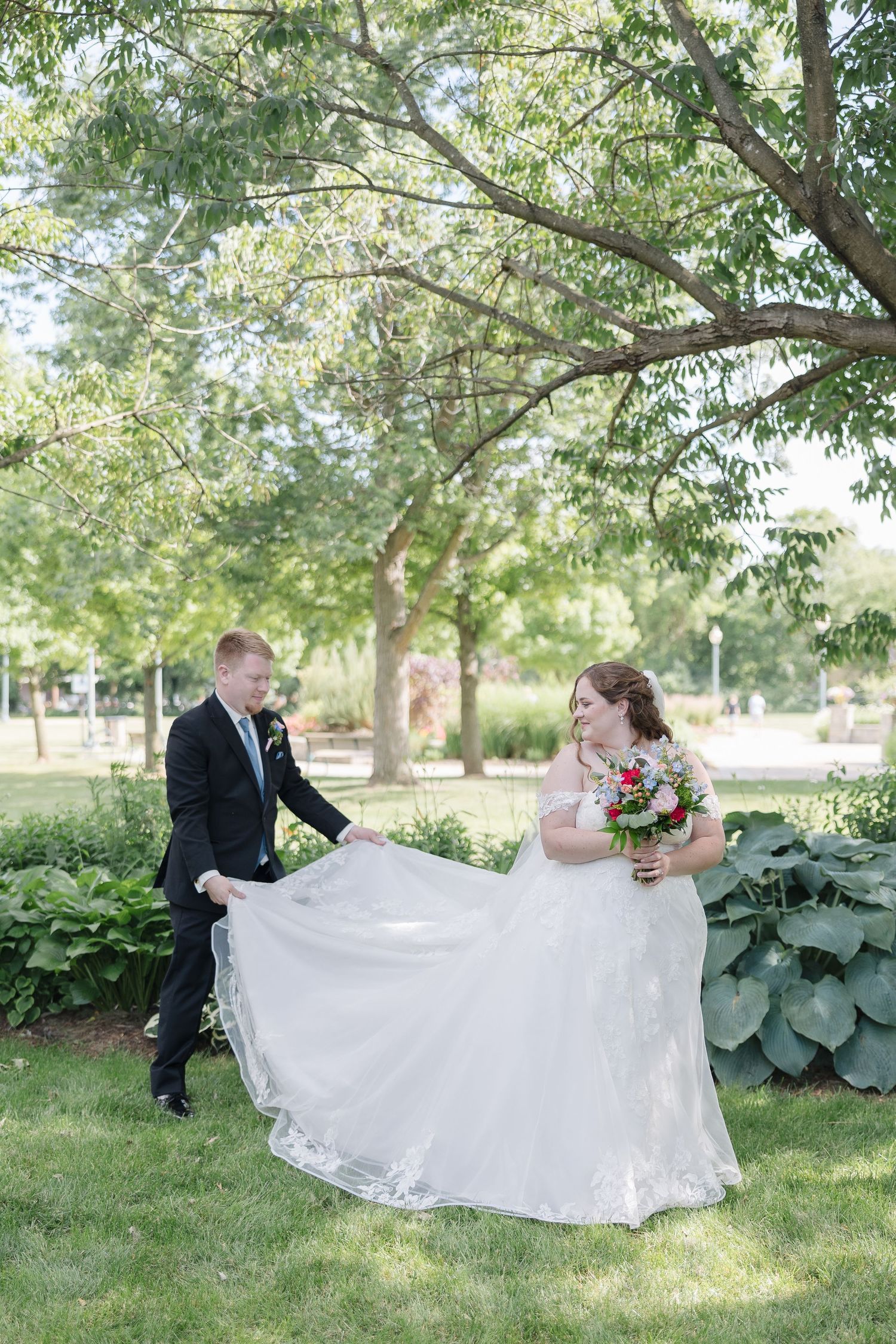 Wedding couple playing with bride's dress in garden setting under green trees.
