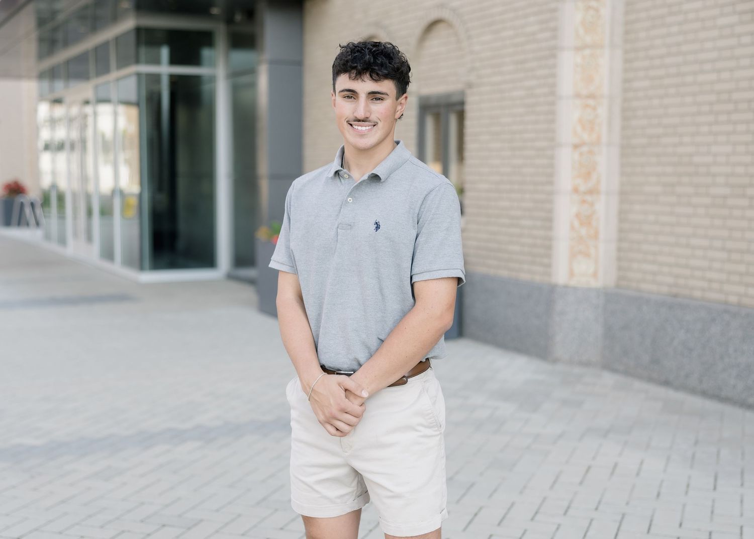 A person in a gray polo shirt and white shorts stands casually in front of a beige brick wall.