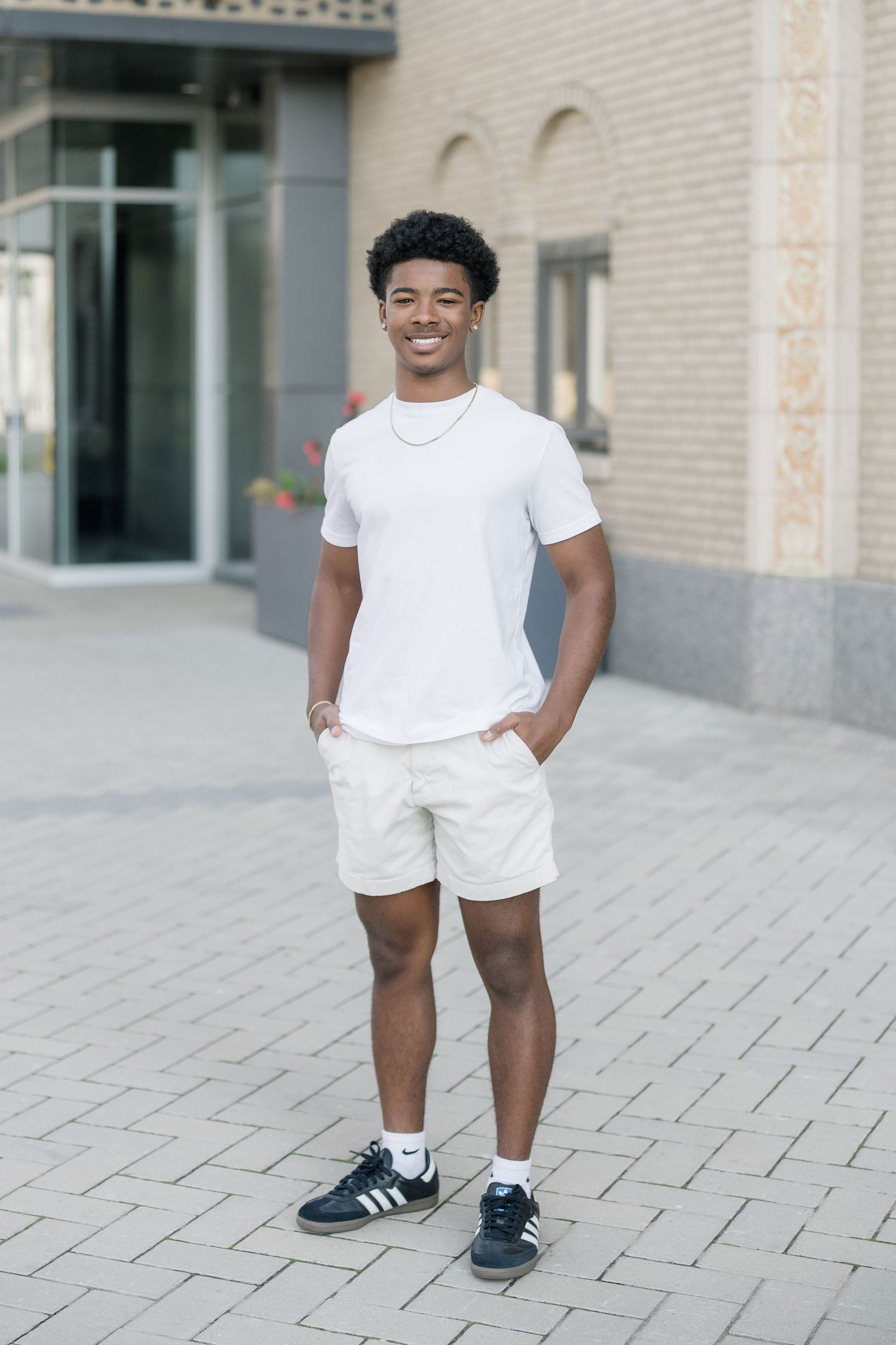 A person wearing a white t-shirt, khaki shorts and Adidas sneakers stands smiling on a brick paved sidewalk.