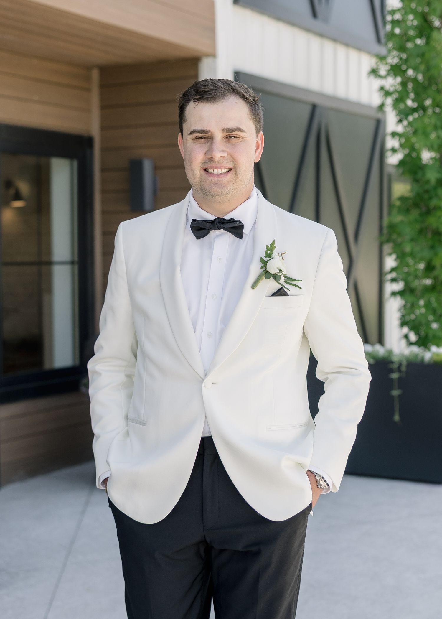 Groom in white tuxedo jacket with black bow tie and floral boutonniere smiling outdoors at wedding venue.