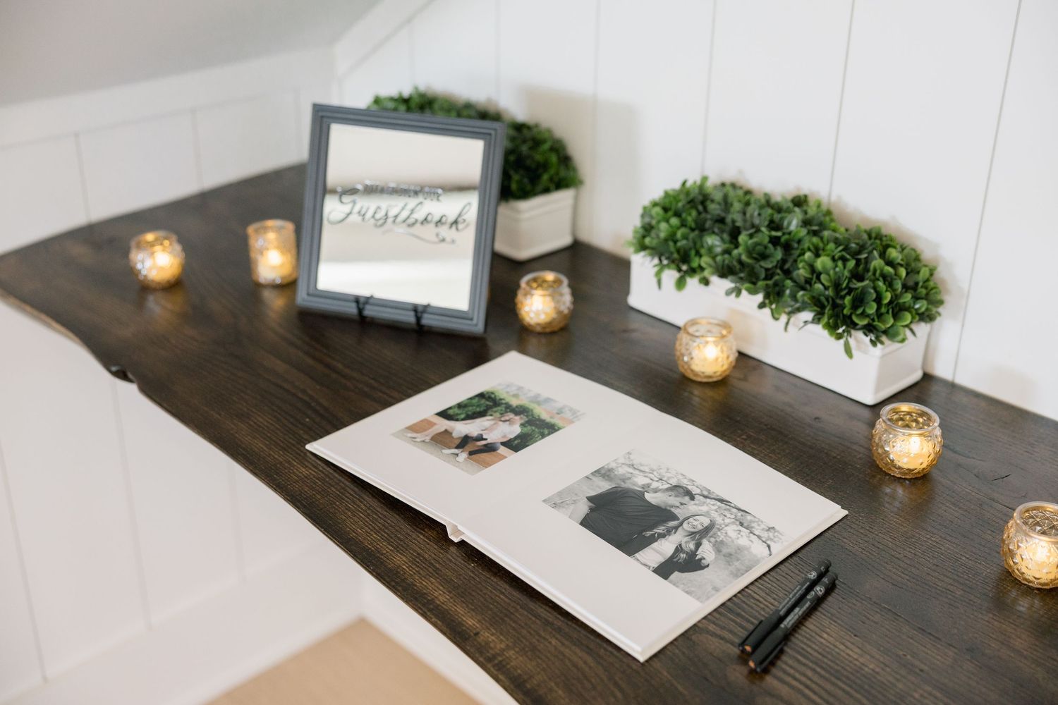 Wedding guest book display on dark wood table with candles, greenery, and framed sign.