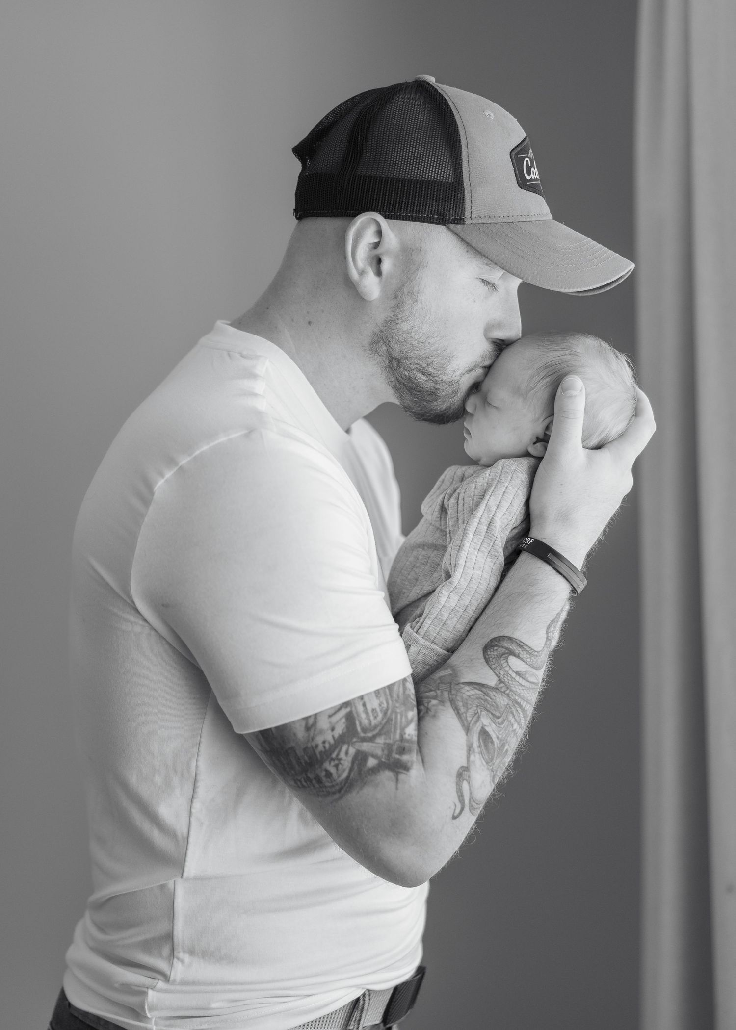A tender black and white portrait of a parent wearing a baseball cap and white t-shirt holding a newborn baby.